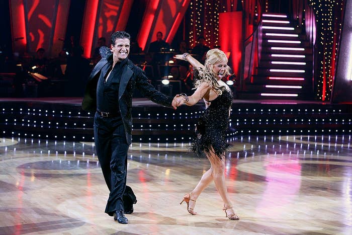 A dance duo performs energetically on a lit stage; the man in a sleek dark suit, the woman in a sparkling fringed dress