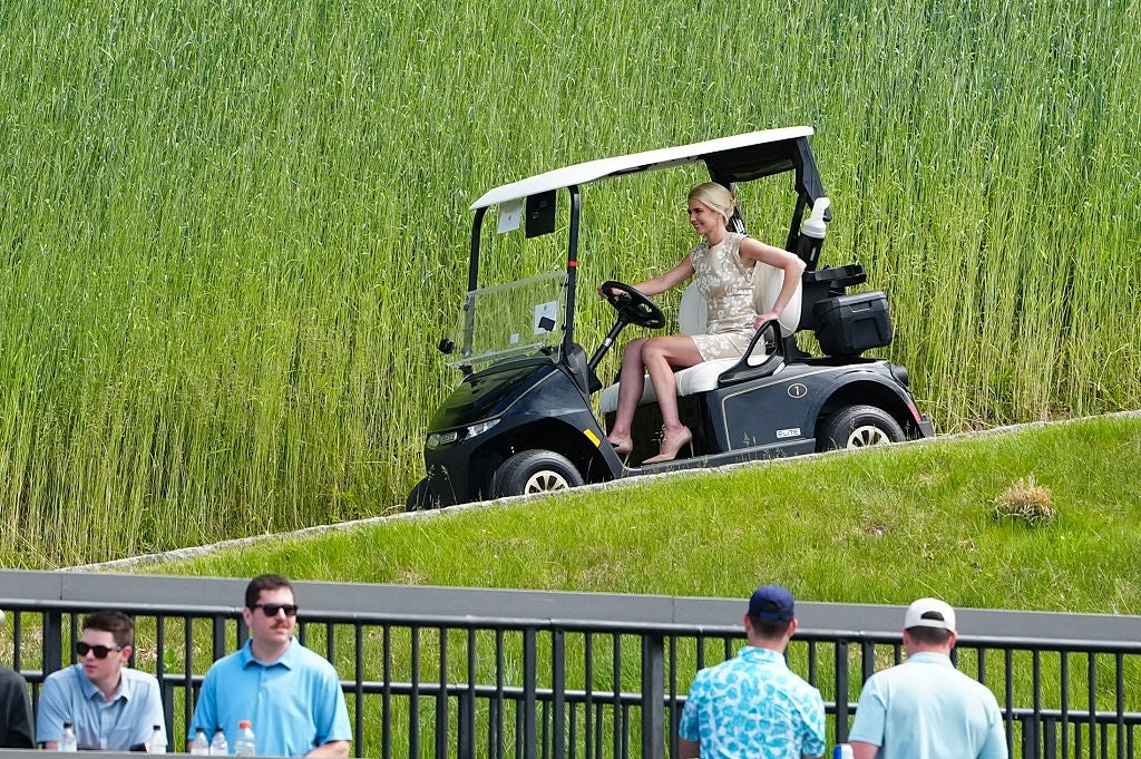 Person i kort kjole kjører en golfbil på en gressbakke mens andre ser nedenfra i nærheten av et gjerde