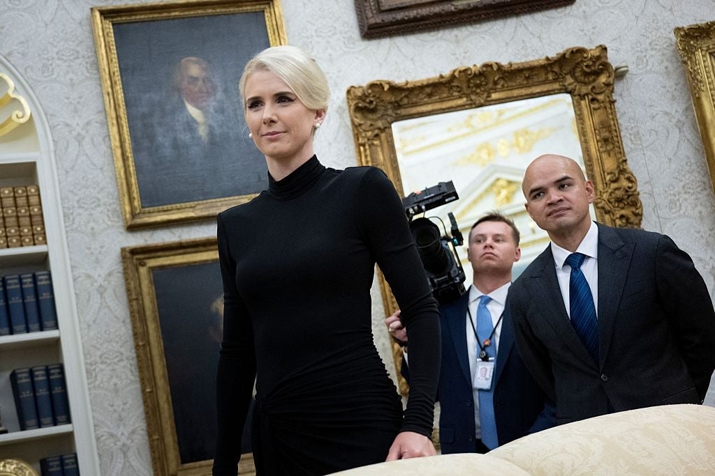 En person i en formell svart kjole går selvsikkert i et rom med innrammede portretter, etterfulgt av to passende personer, den ene holder et kamera