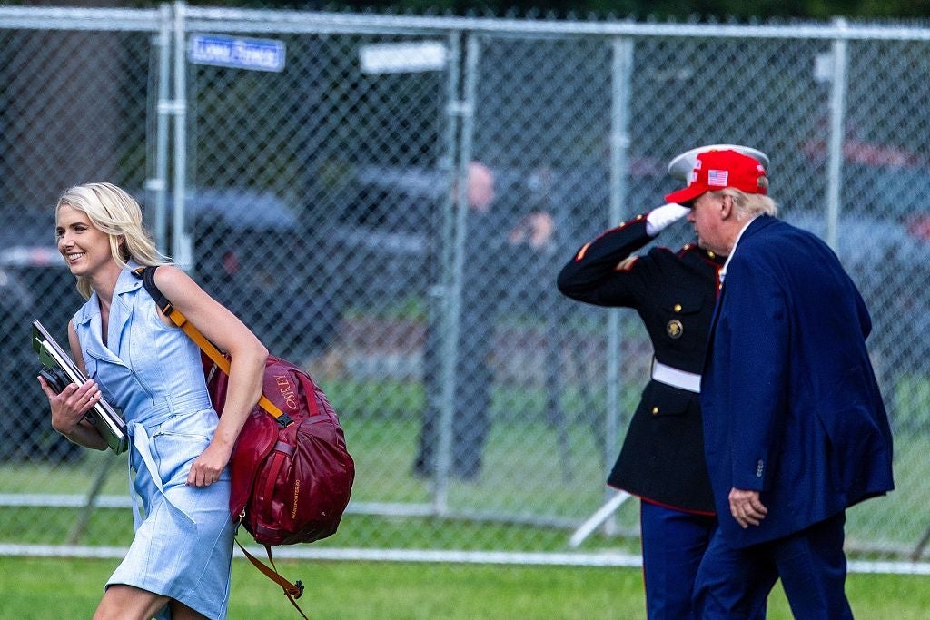 Kvinne som smiler, bærer bøker og en stor bag, går forbi en hilsende marinesoldat og en mann i dress med rød lue
