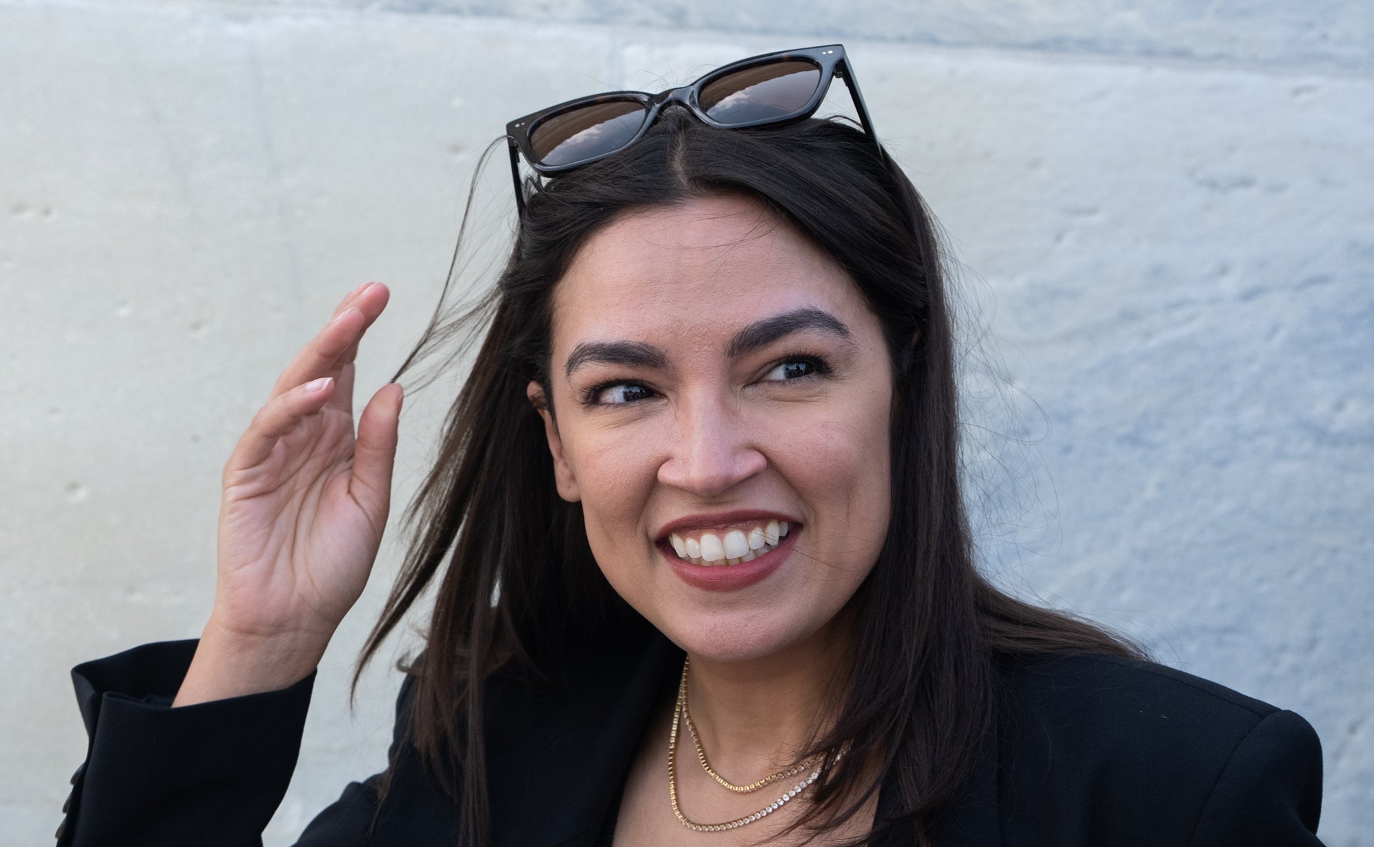 Alexandria Ocasio-Cortez iført solbriller på hodet og en stilig blazer, smiler selvsikkert med hånden hevet nær ansiktet