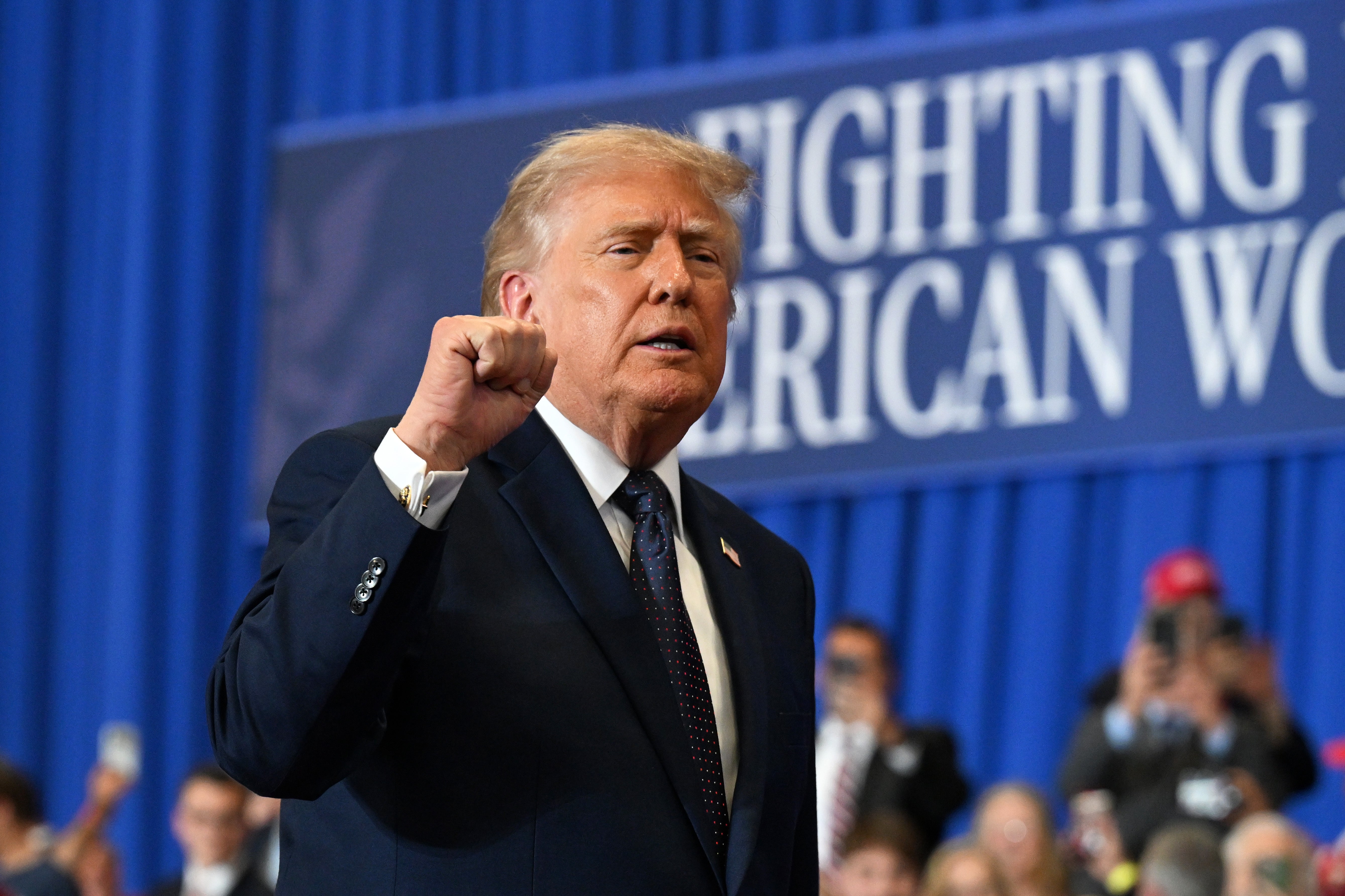 En person i dress hever knyttneven ved et offentlig arrangement, og står foran en "Fighting for the American Worker" skilt med folk i bakgrunnen