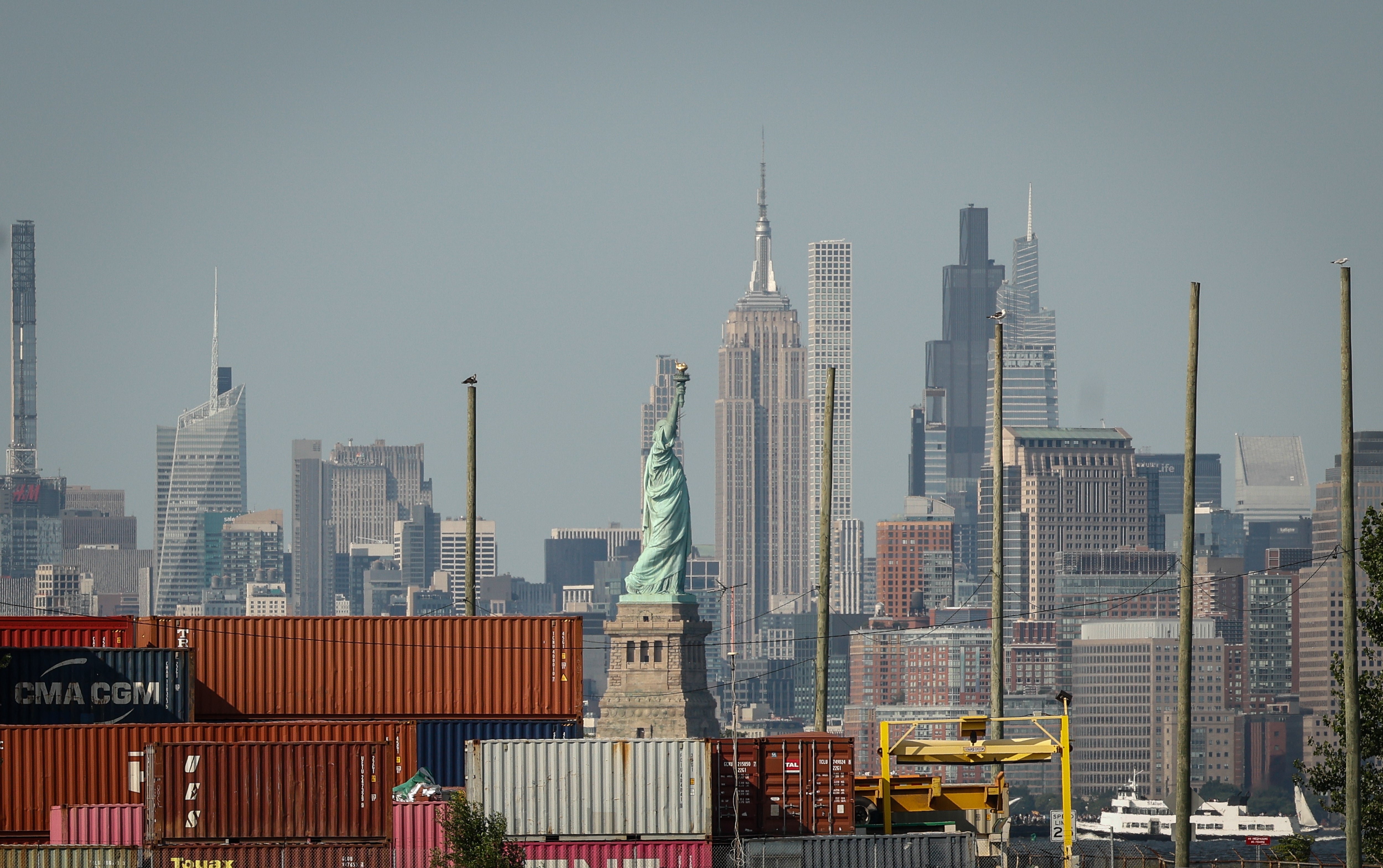 Frihetsgudinnen står blant fraktcontainere, New York Citys skyline er synlig i bakgrunnen