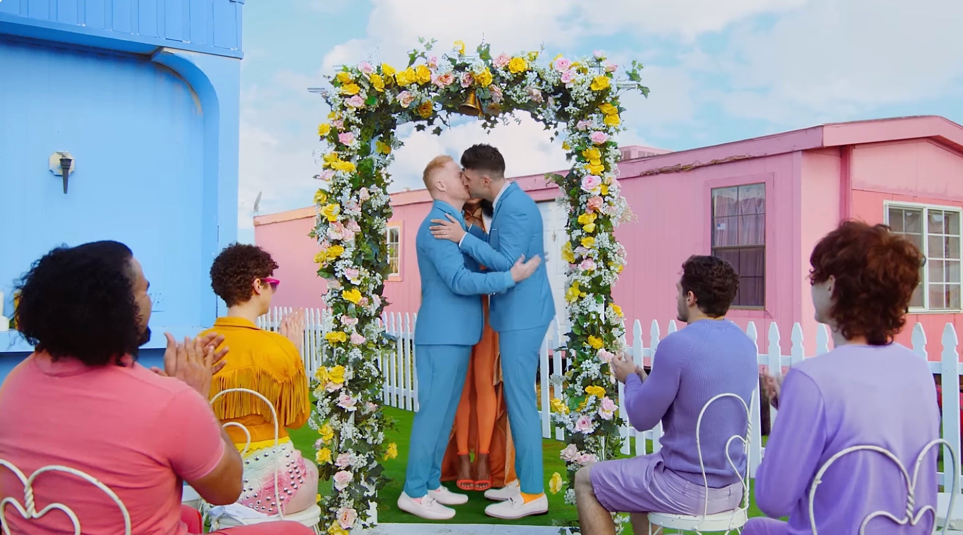 Taylor Swift's "You Need To Calm Down" music video, featuring two men in matching suits kiss under a floral altar at an outdoor wedding ceremony.