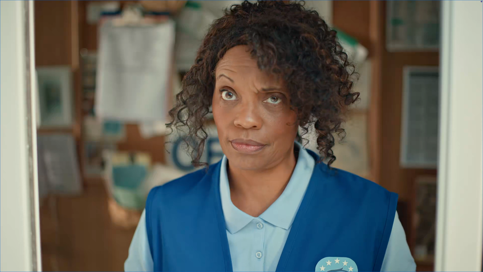Person with curly hair gives a skeptical look, wearing a blue collared shirt and vest with a star emblem. Background has papers and a soft-focus effect