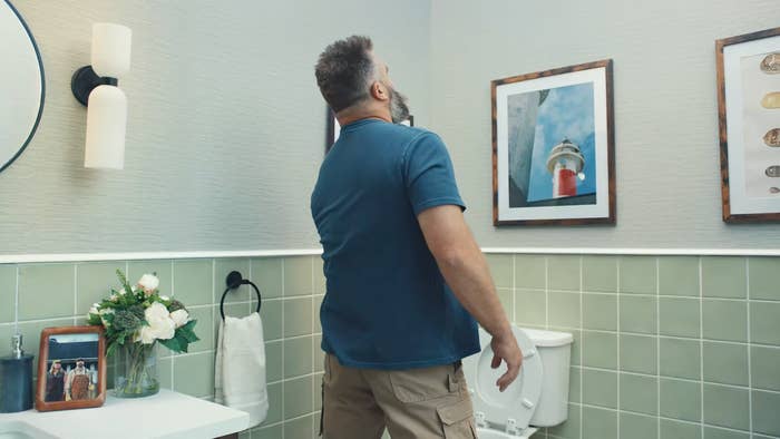 A man in a bathroom looks up with a surprised expression. The room has framed art and green-tiled walls