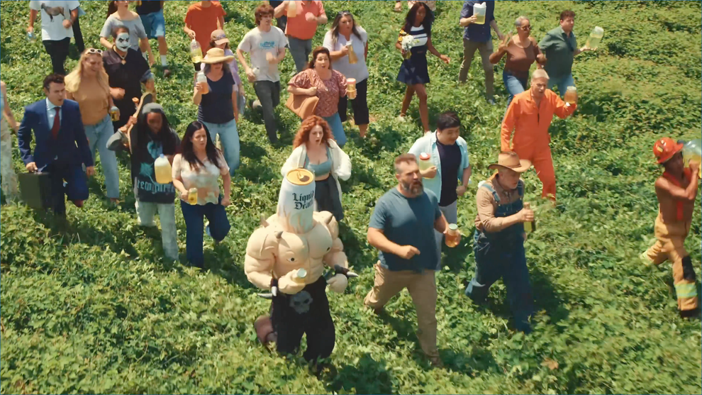 A group of people in various costumes and attire, including a person with a large can on their head, walk energetically through a field