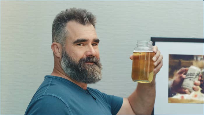 A man with a beard smiles slightly while holding a jar of liquid in a casual setting. A framed photo is visible in the background