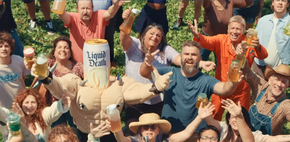 Group holding mugs in a garden, led by a person in a can costume. Text: "We want your pee. Drink Liquid Death Energy and Garage Beer."