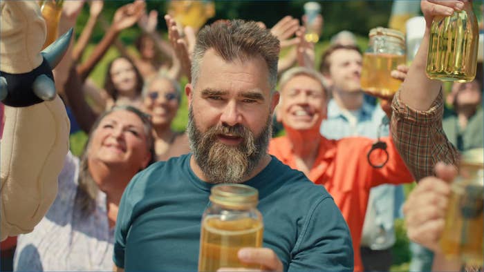 A bearded man holds a jar of honey while others cheerfully raise jars in the background