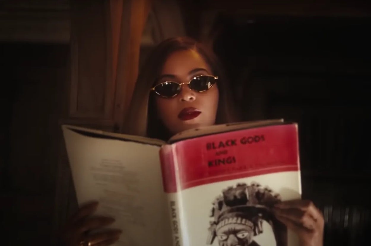 Person reading a large book titled "Black Gods and Kings" while wearing sunglasses indoors