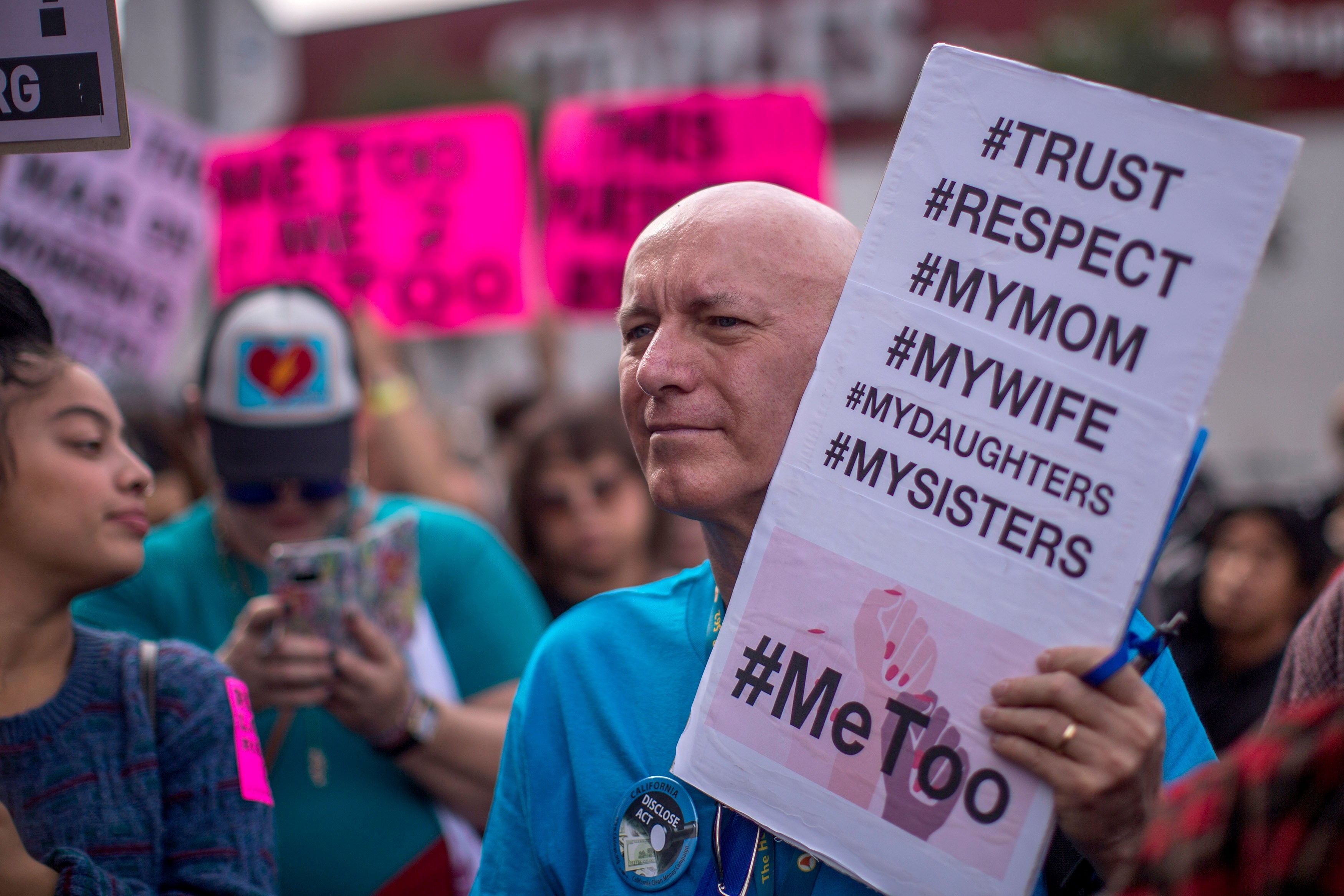 A man holds #MeToo sign with hashtags #Trust, #Respect, #MyMom, #MyWife, #MyDaughters, and #MySisters at a MeToo protest.