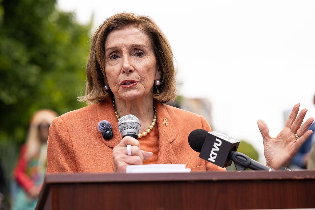Nancy Pelosi snakker på et podium utendørs, holder en mikrofon, med flere mikrofoner foran seg