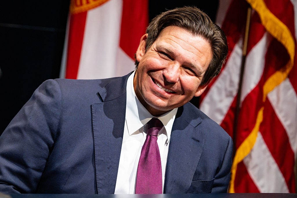 Smilende Ron DeSantis i dress og slips står foran amerikanske flagg