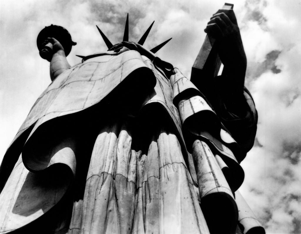 Low-angle view of the Statue of Liberty with a cloudy sky background. The focus is on the structure's impressive size and detail