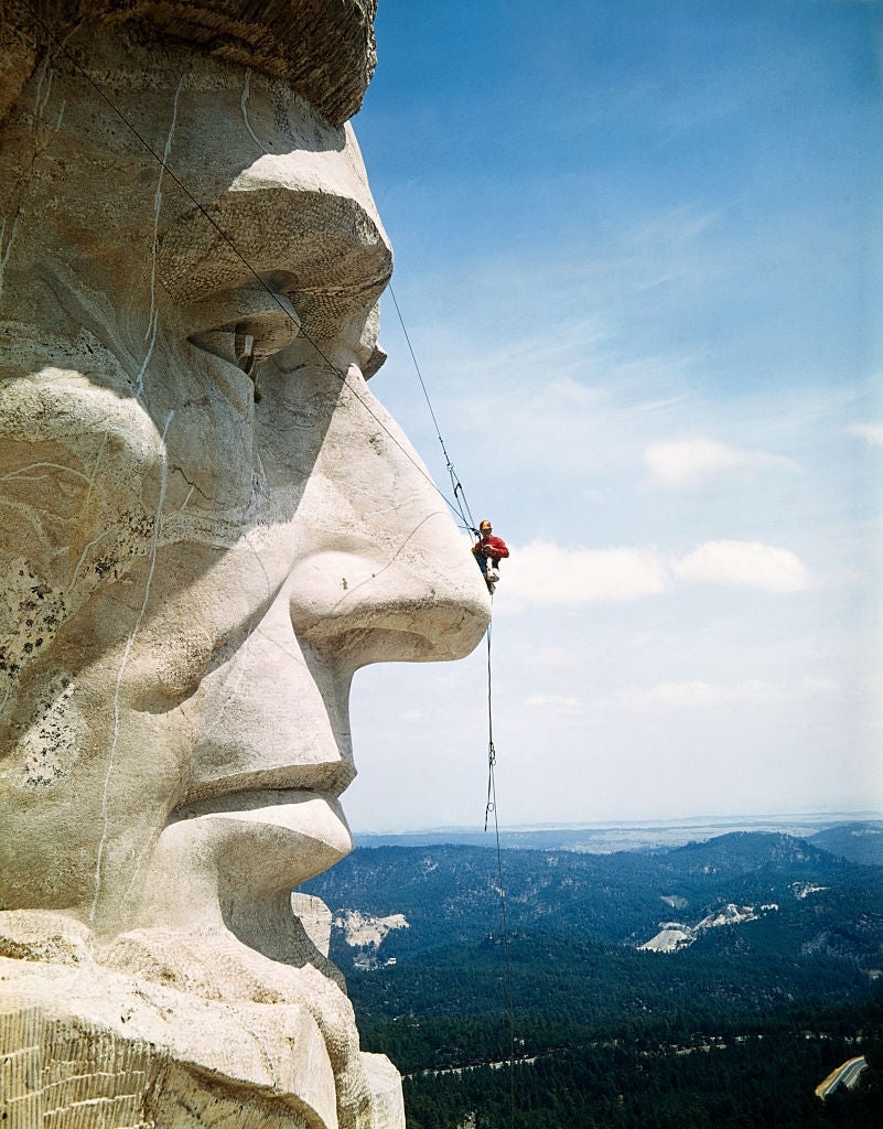 A climber hangs from ropes on the nose of a large carved stone face on Mount Rushmore, with forested hills in the background