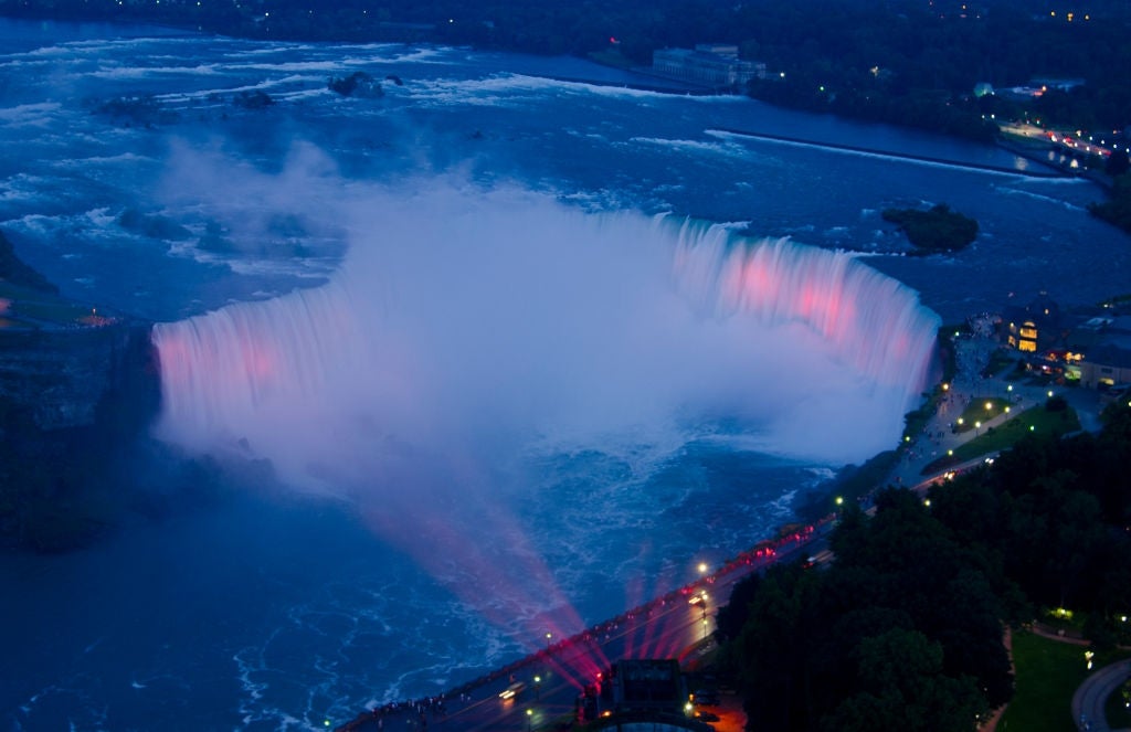 Aerial view of Niagara Falls at night, with spotlights casting colorful lights on the cascading water and surrounding landscape