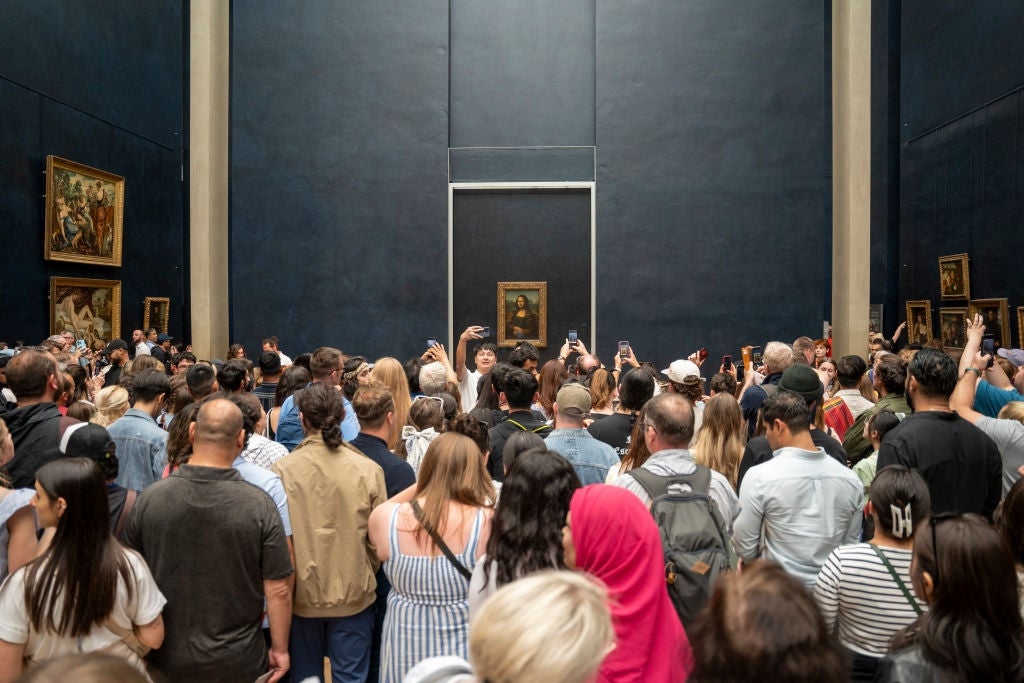 A large crowd gathers in a museum gallery, many taking photos. The Mona Lisa is prominently displayed on the wall