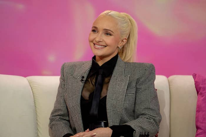 Person sitting on a sofa, smiling, wearing a checkered blazer over a sheer top with a tie