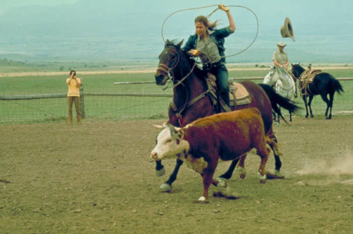 Jane Fonda rides horseback and lassos a calf in a rodeo arena, with spectators in the background. The image is from the film "Comes a Horseman"