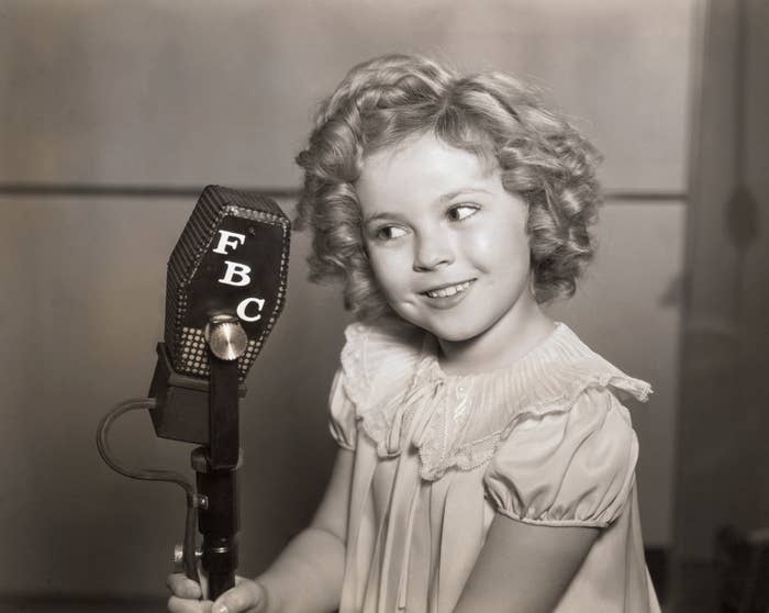 Shirley Temple wears a frilly dress and speaks into an old-fashioned microphone labeled "FBC."