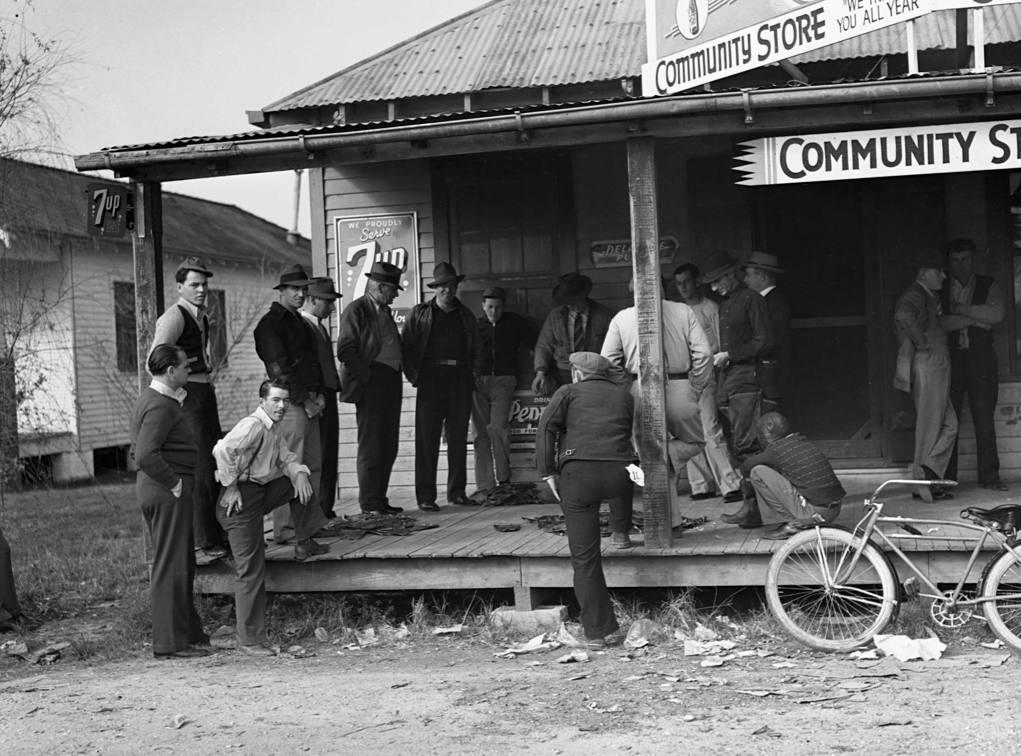 People gathered on a wooden porch of a community store, with a bicycle nearby. Some lean against posts, engaging in conversations