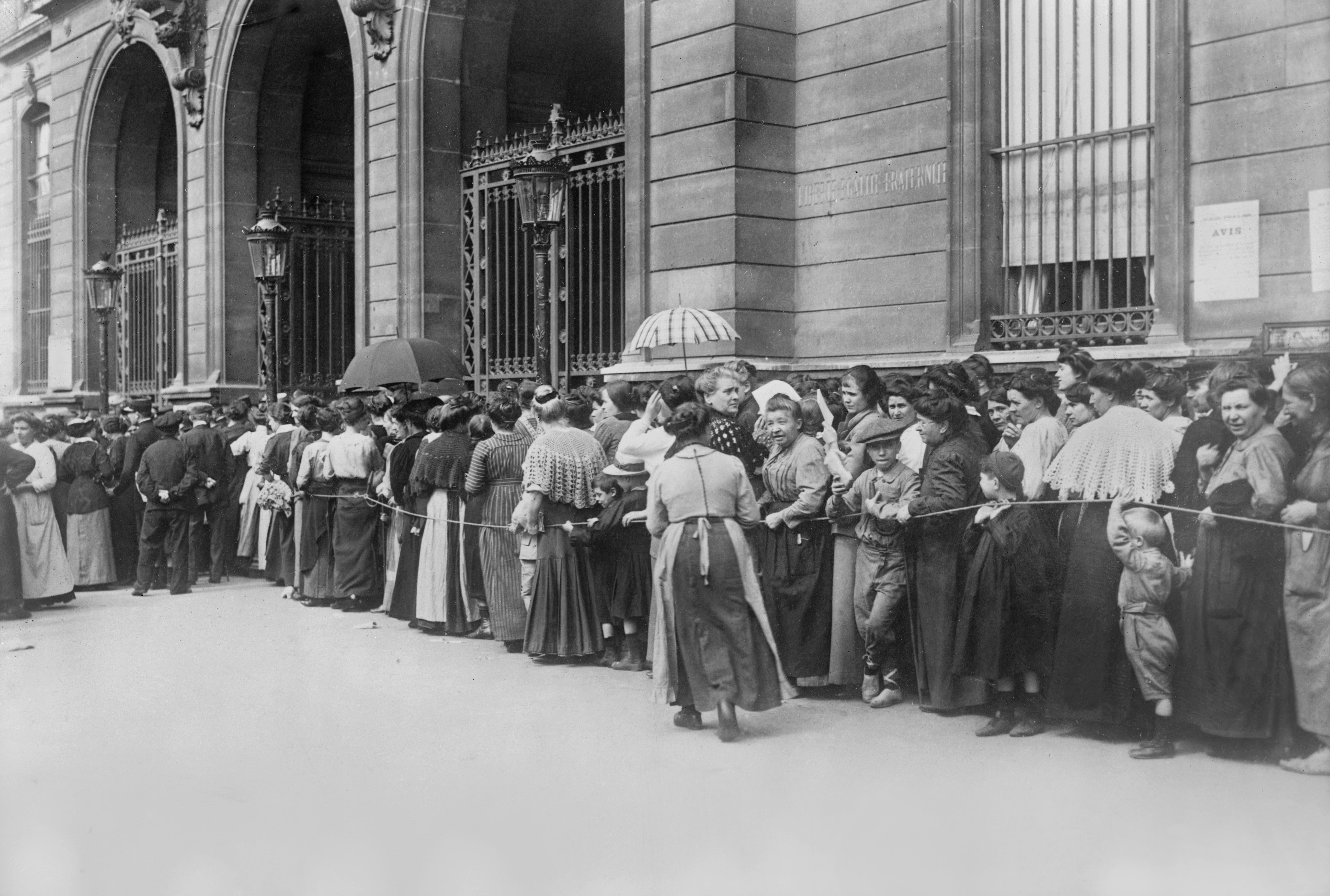 A long line of people wait outside a building, some holding umbrellas