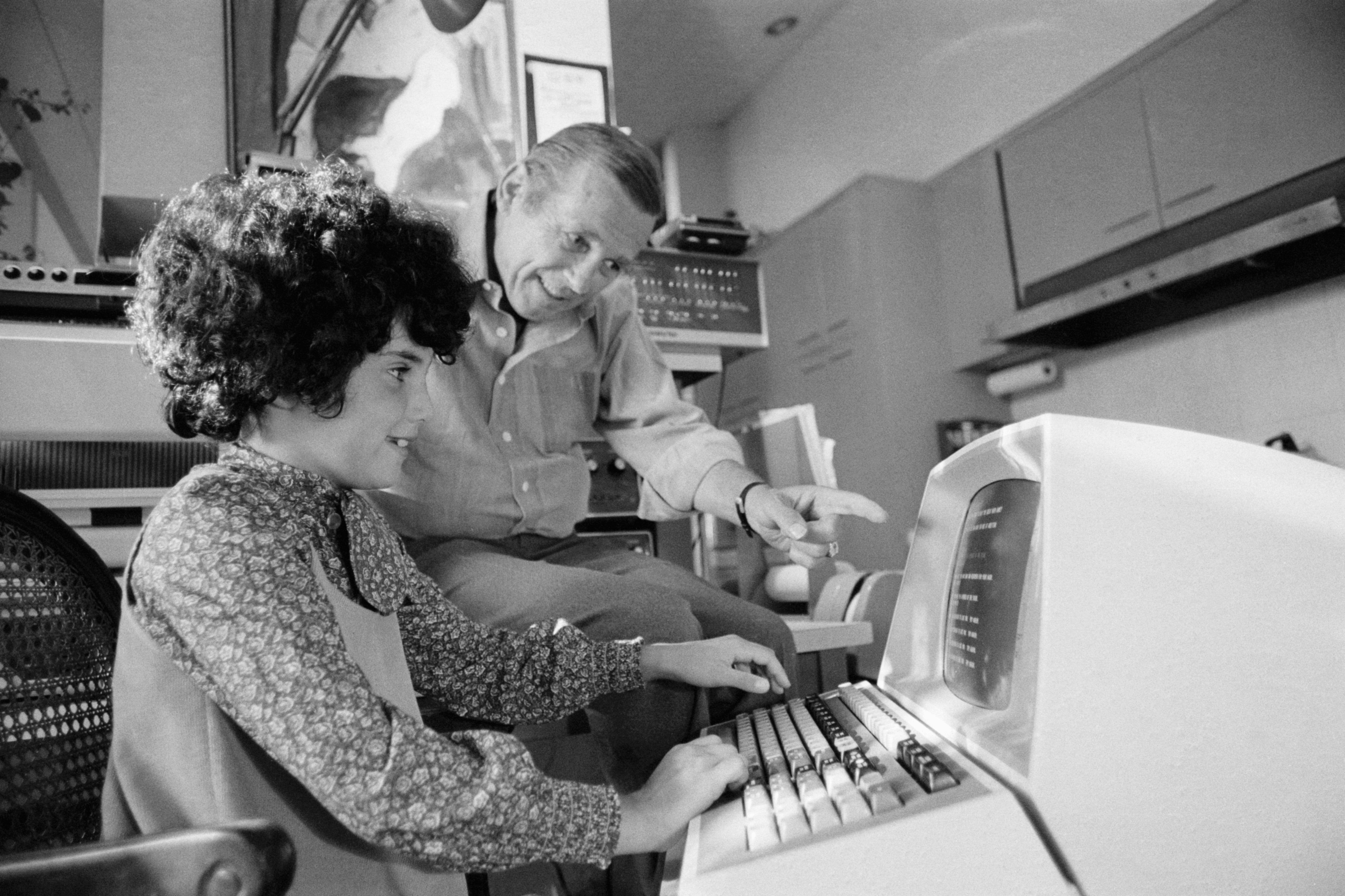 A young child types on an early computer keyboard, guided by their father pointing at the screen, in a retro office setting
