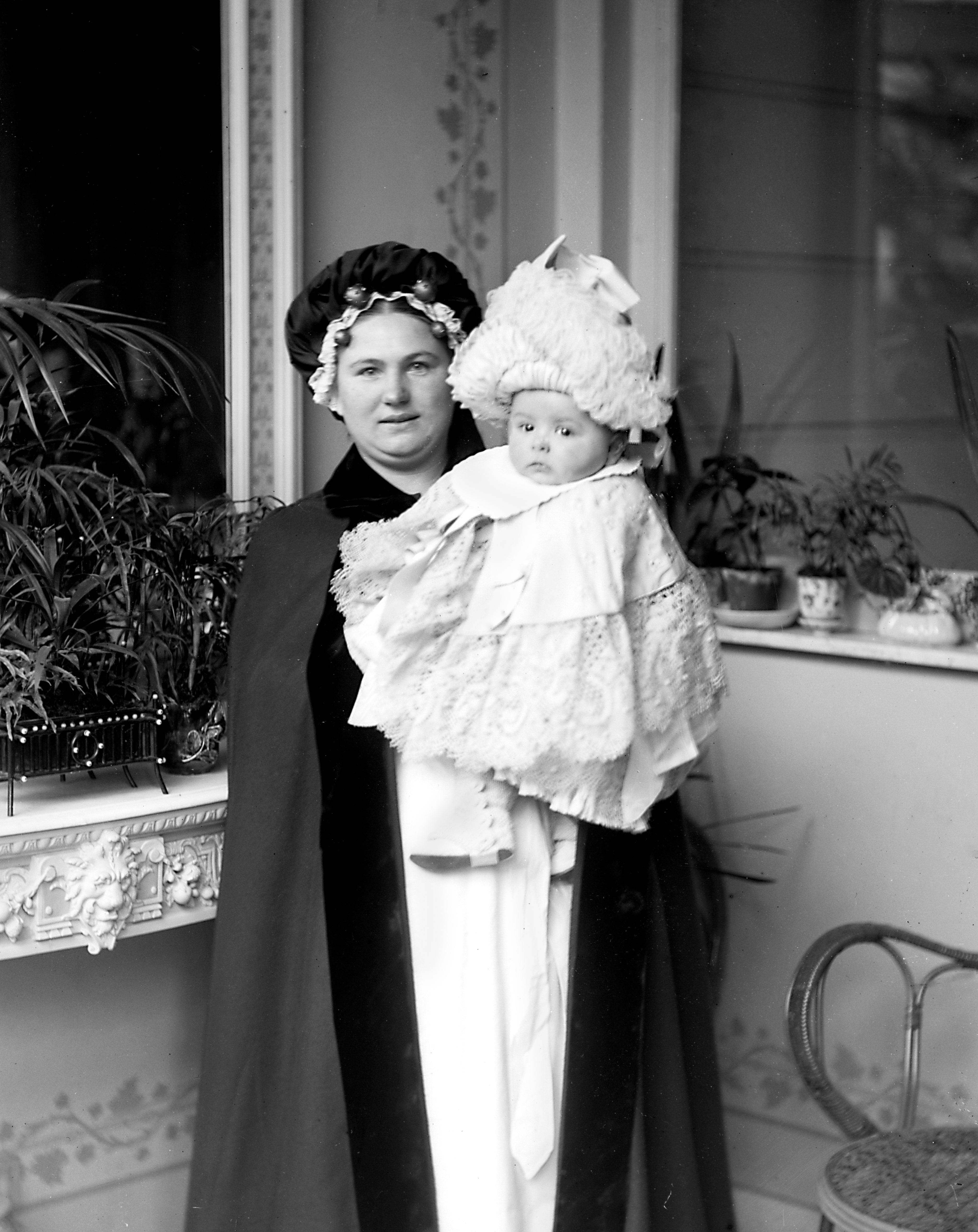 A woman in fancy period clothing holds a baby wrapped in lace, standing on a decorated veranda