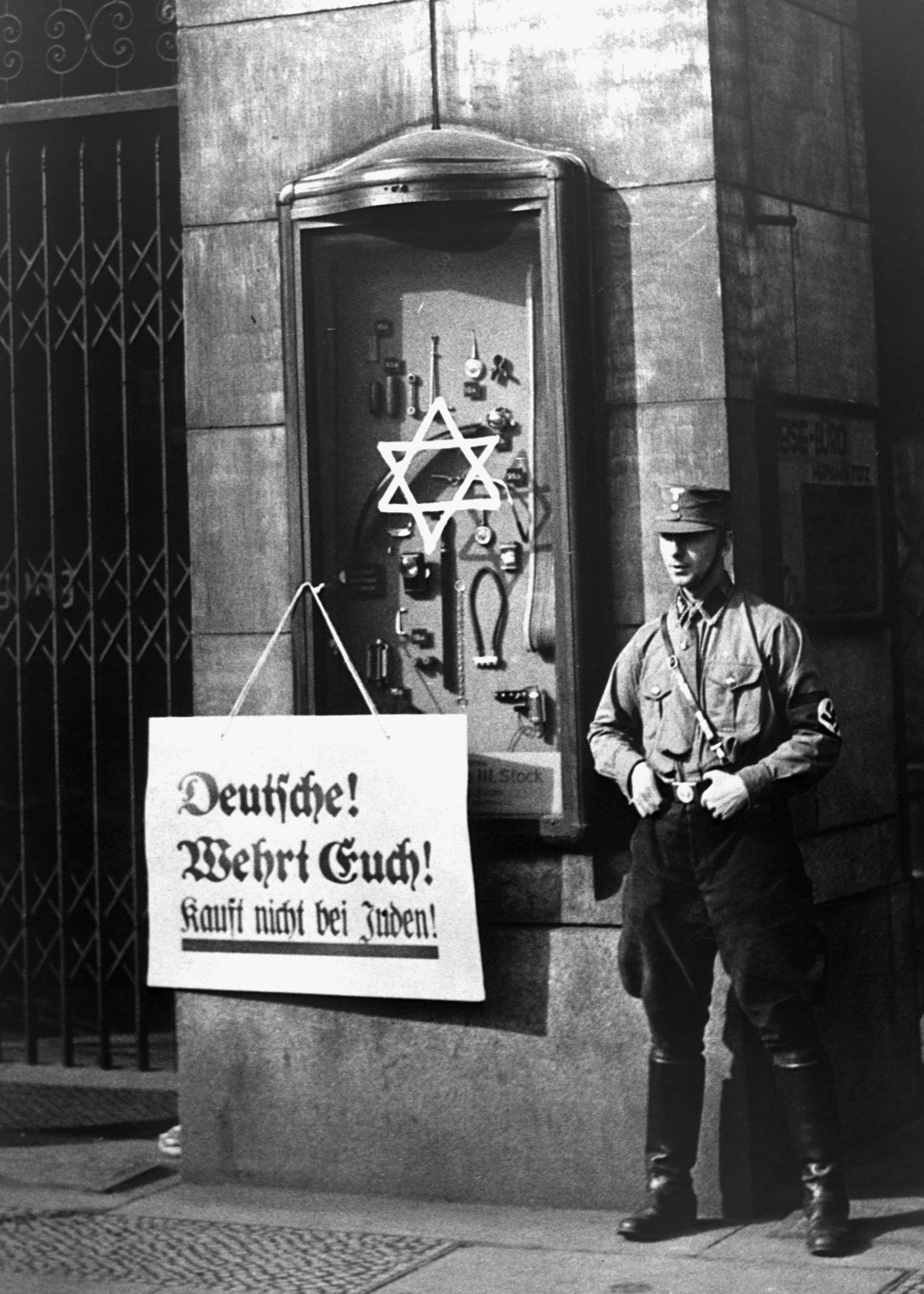 A man in uniform stands beside a storefront with anti-Semitic signage and a Star of David, promoting a Nazi boycott of Jewish businesses