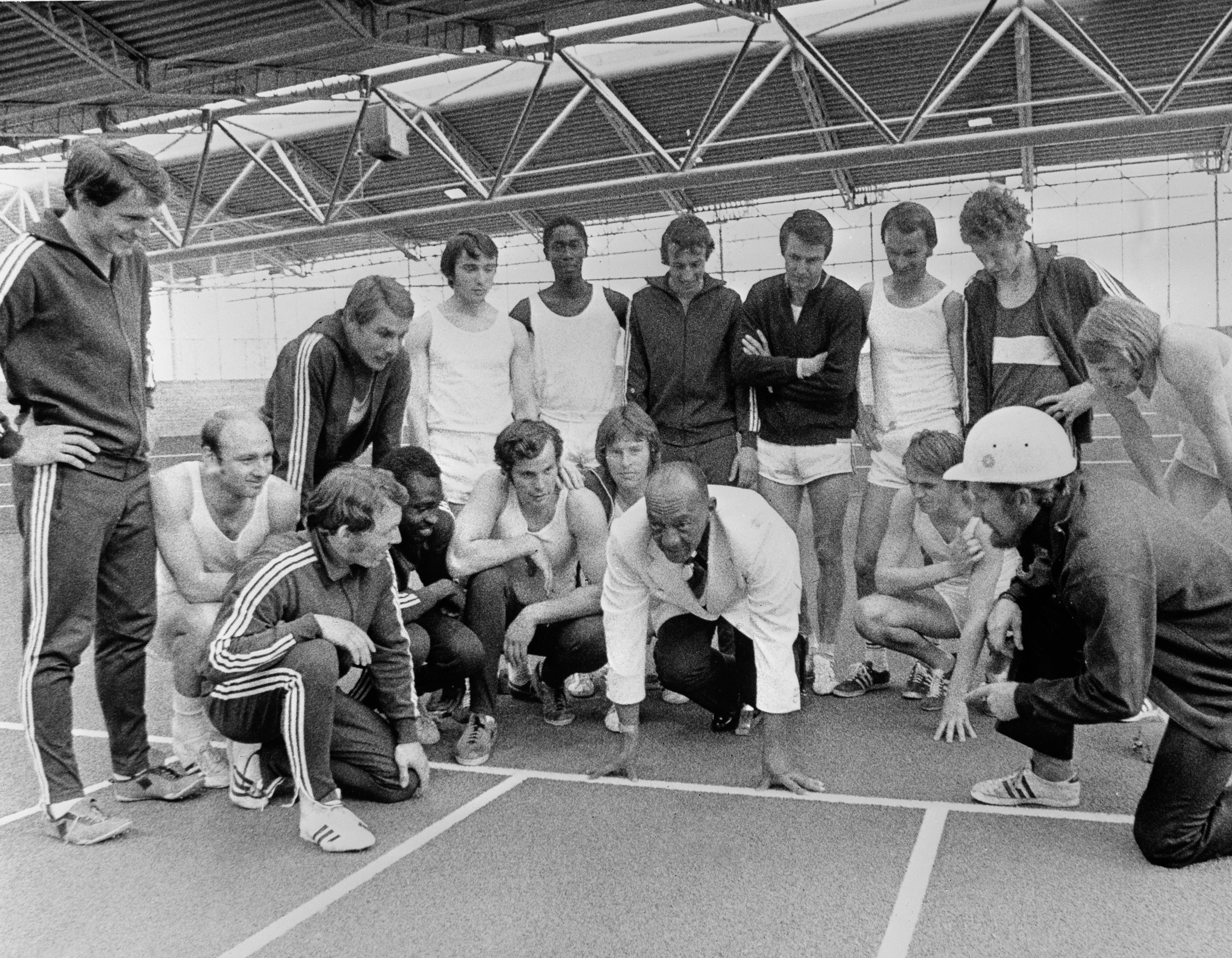 Group of athletes in a gym listen to Jesse Owens demonstrating a technique on the track. Some wear tracksuits, others shorts and tank tops