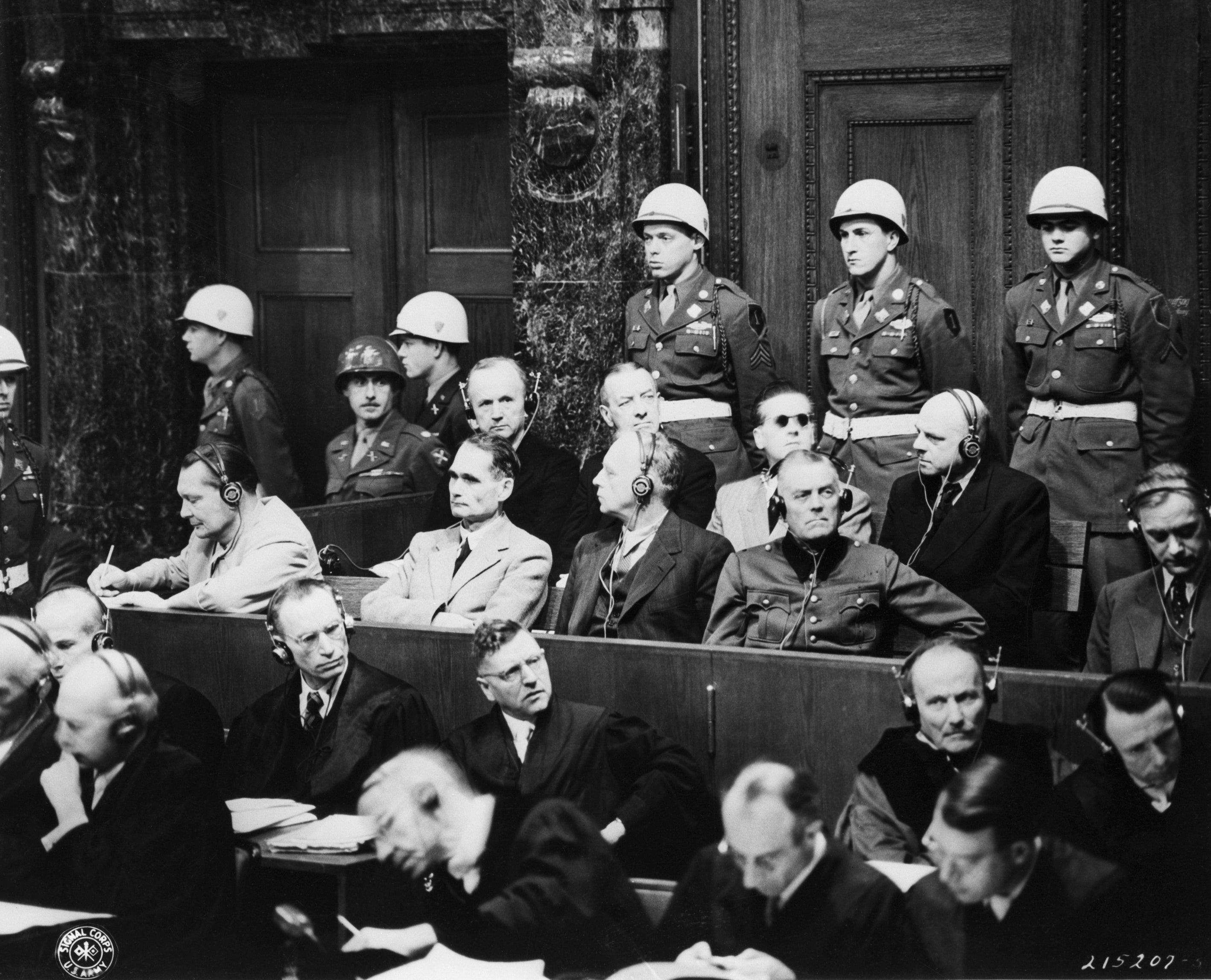 Historical courtroom scene with military officers standing behind suited individuals wearing headphones