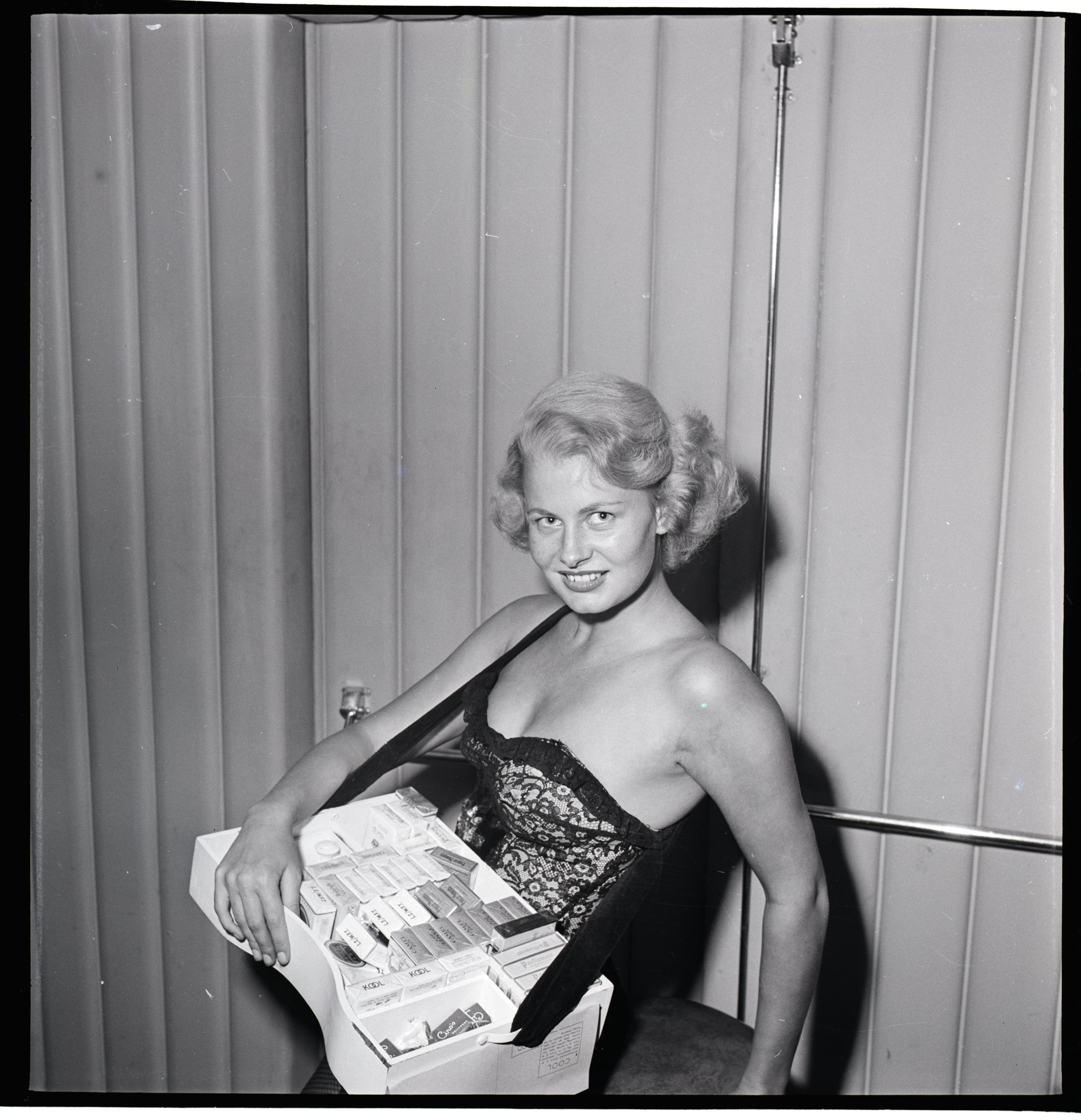 Woman wearing a strapless, lacy corset dress, holding a display of various cigarette boxes, smiling at the camera