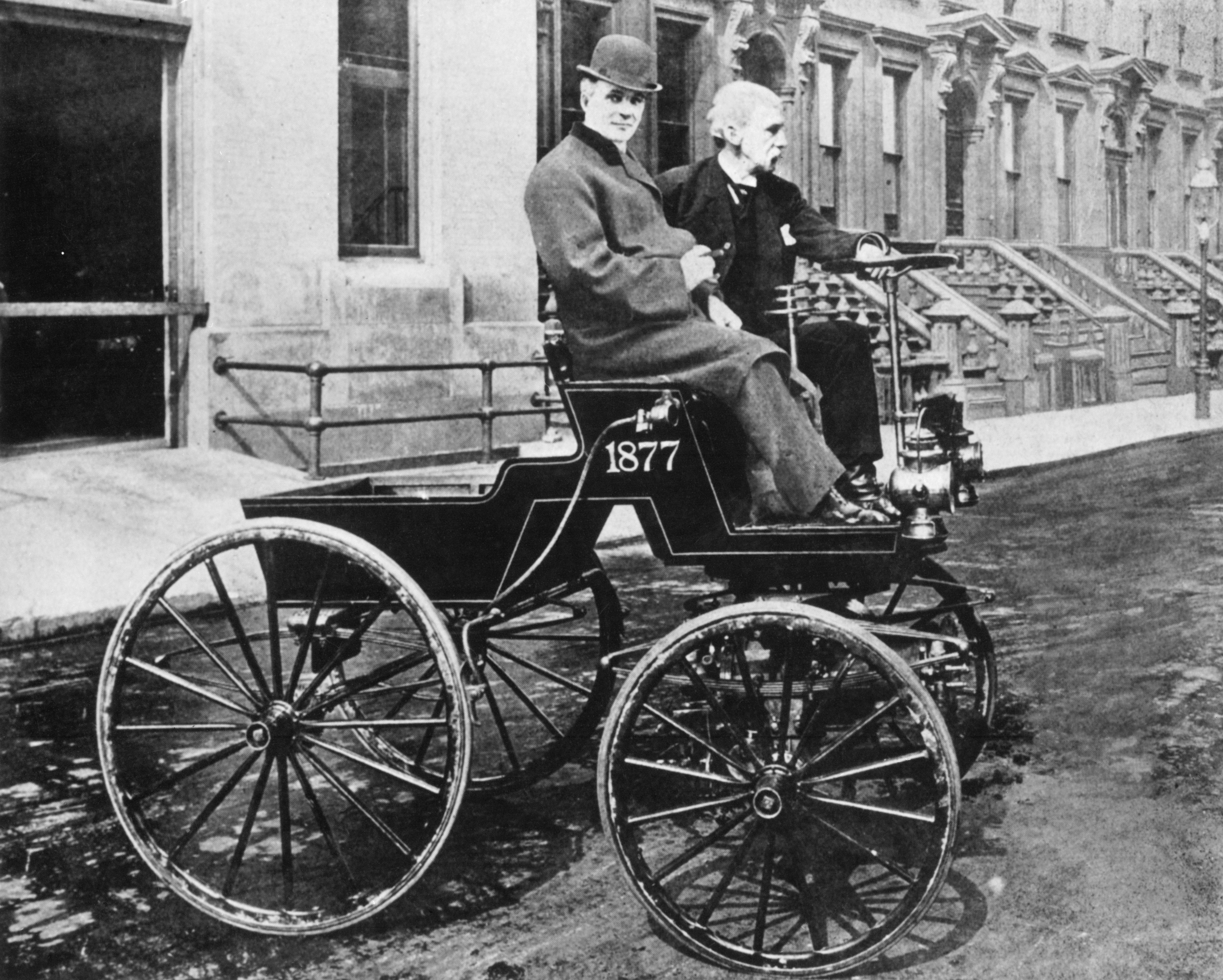 Two men, Henry Ford and George B. Selden, sit in an early motor car from the late 1800s on a city street, wearing period clothing