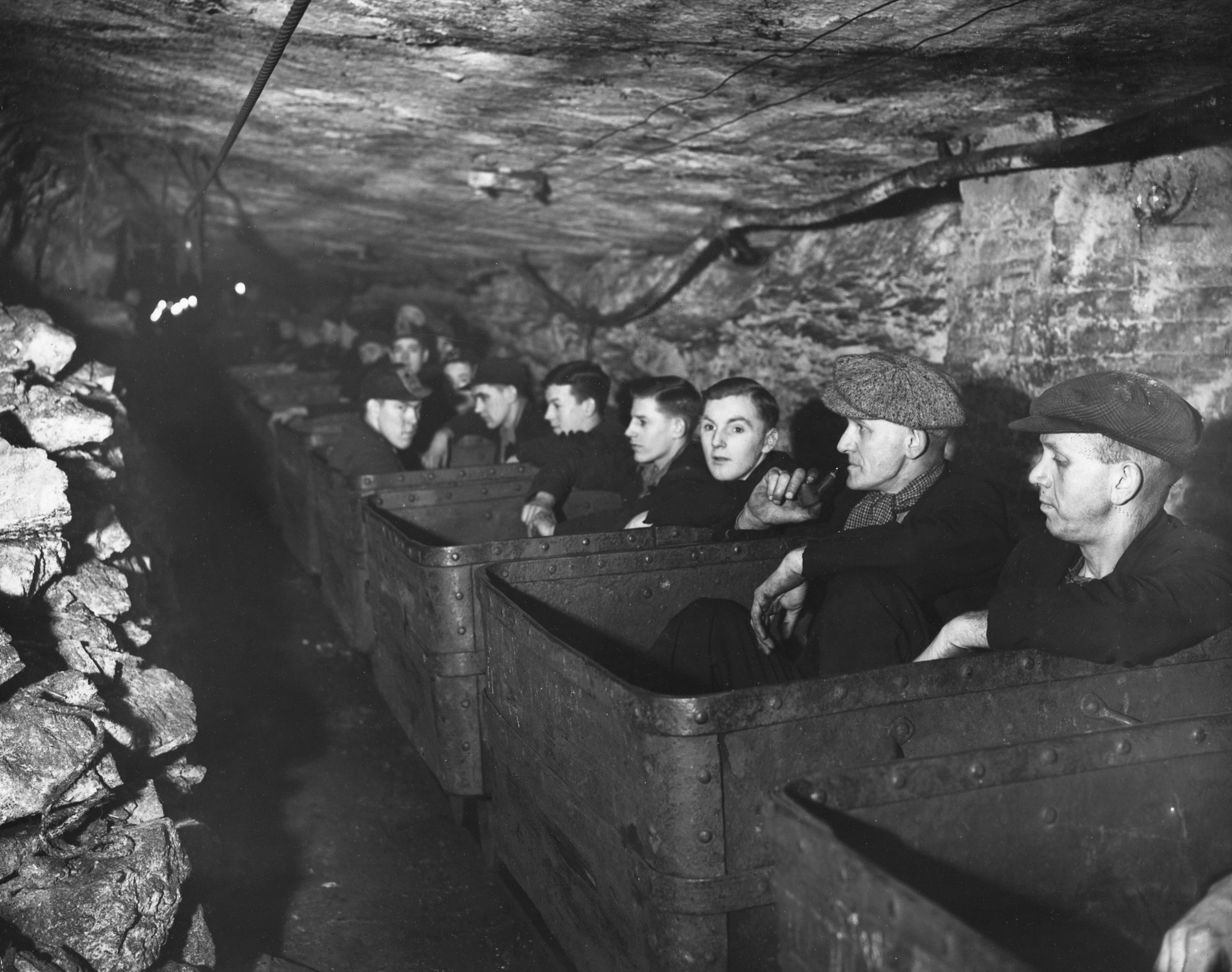 Miners wearing flat caps and work attire sit in carts inside a narrow underground tunnel, ready to begin their shift