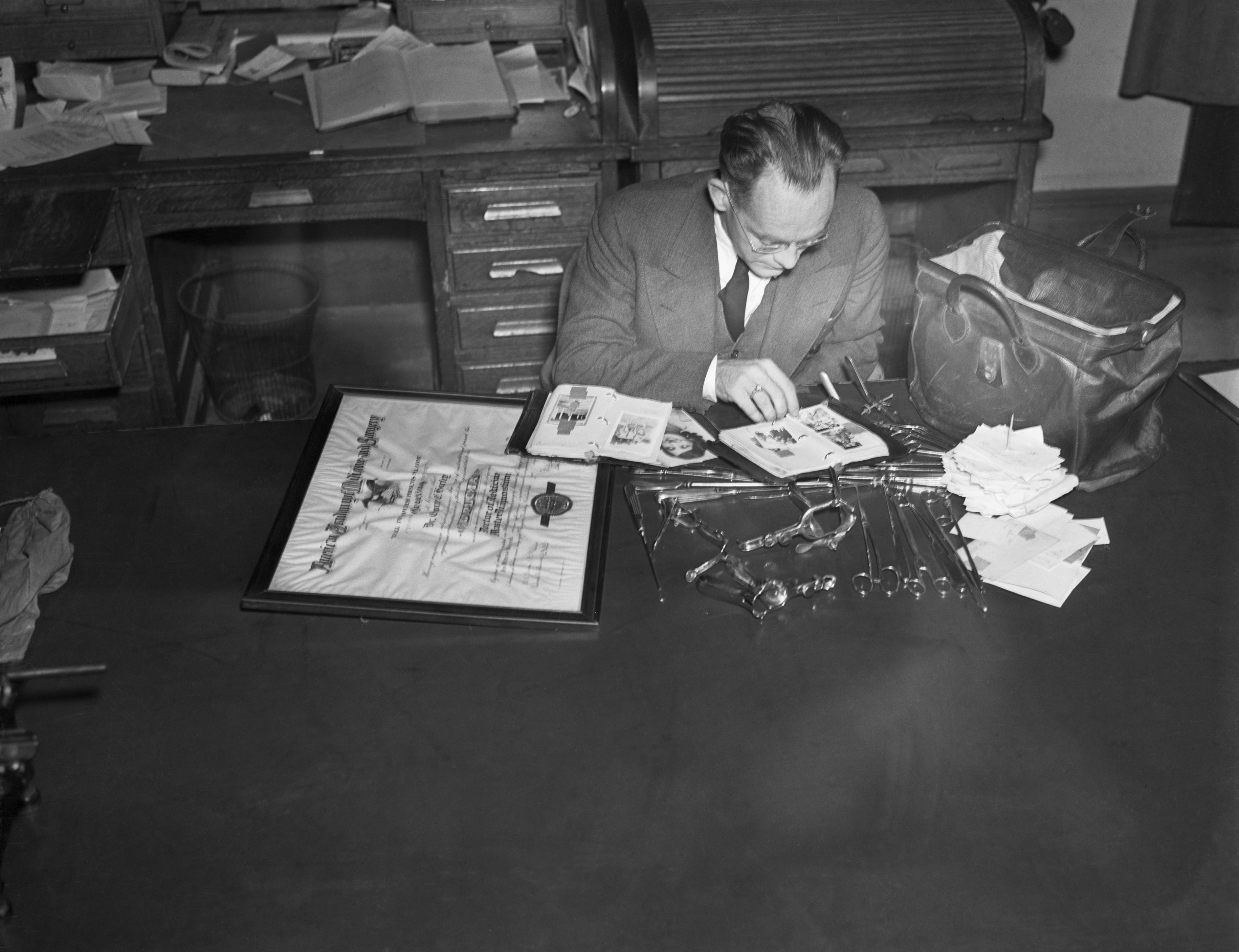 A doctor sits at a desk examining papers, surrounded by scattered papers, stamps, and an open mailbag