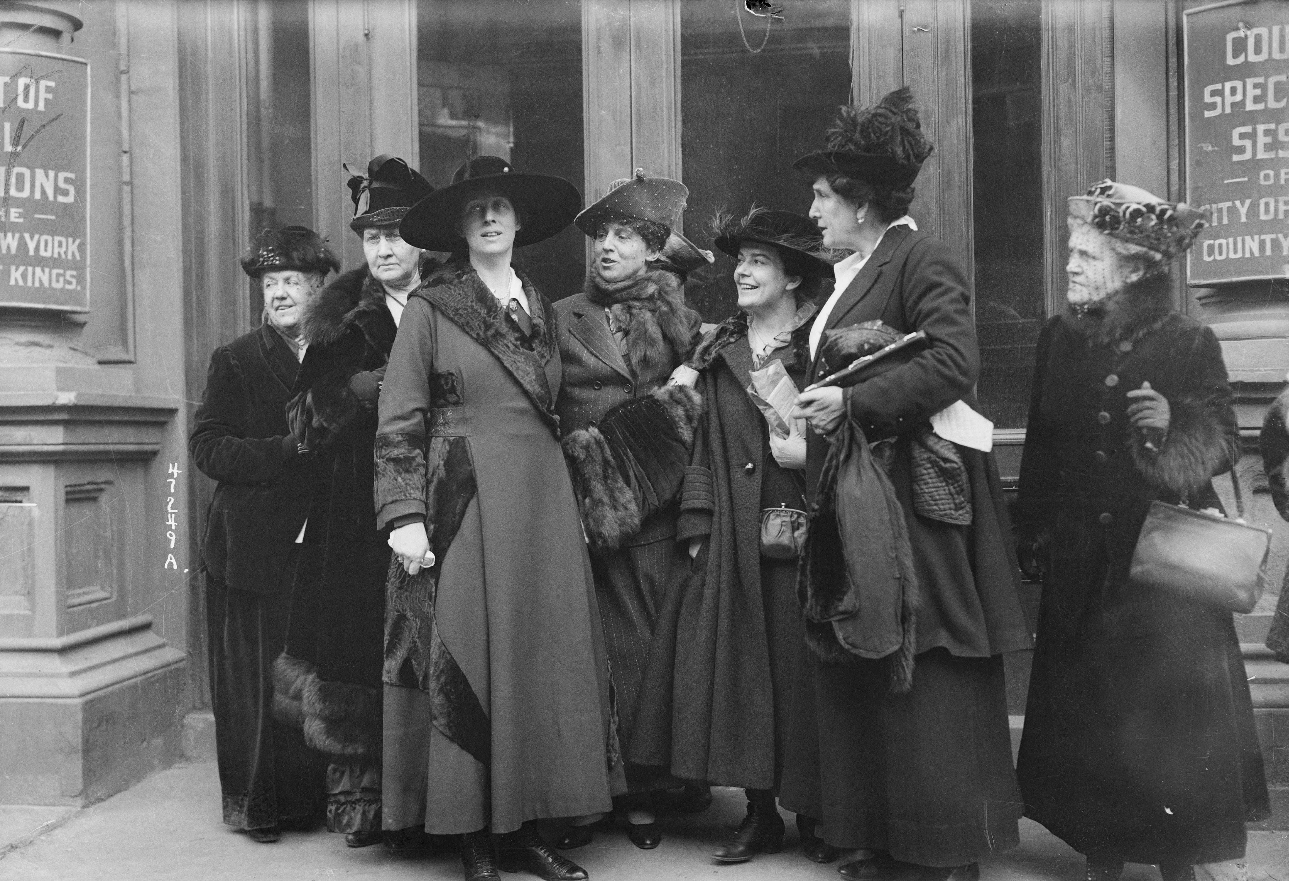 A group of well-dressed women in early 20th-century attire, wearing hats, gather outside a building, engaged in conversation