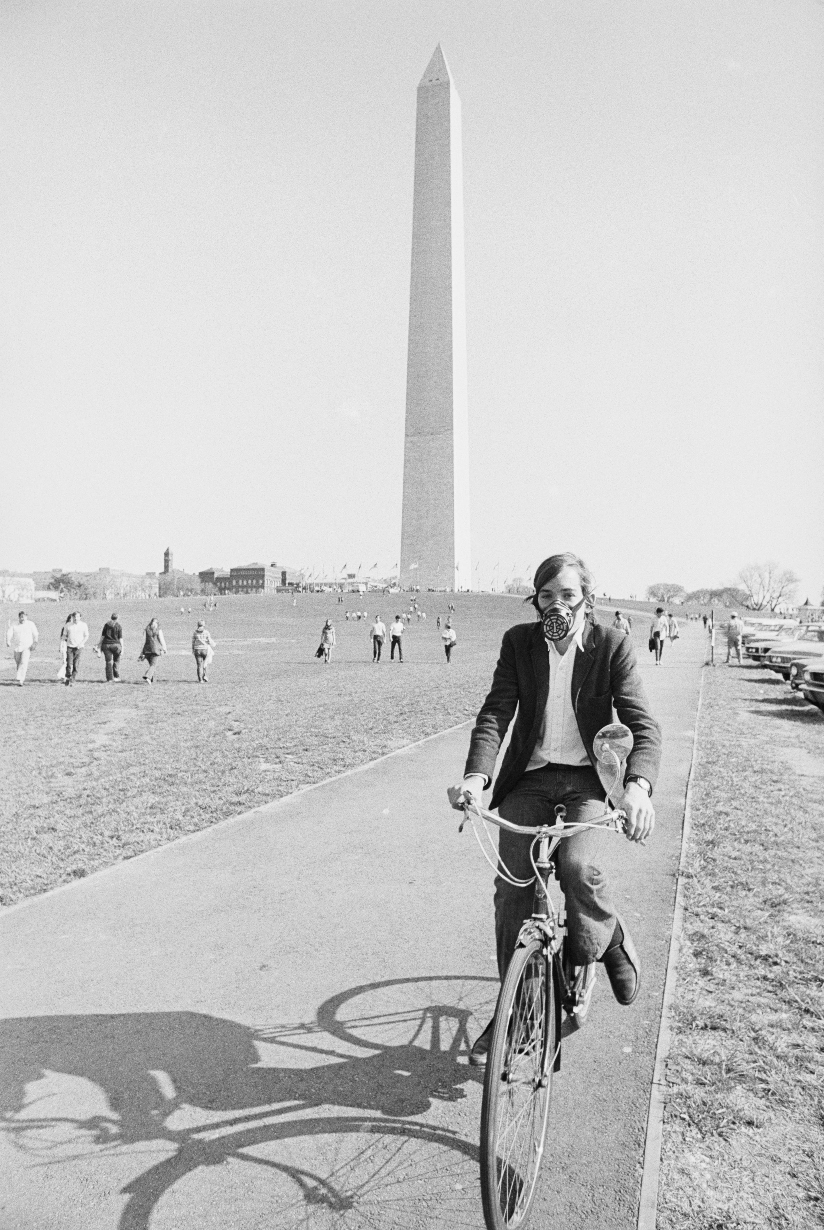 Person cycling on a path near the Washington Monument, with others walking in the background. He is wearing a gas mask.