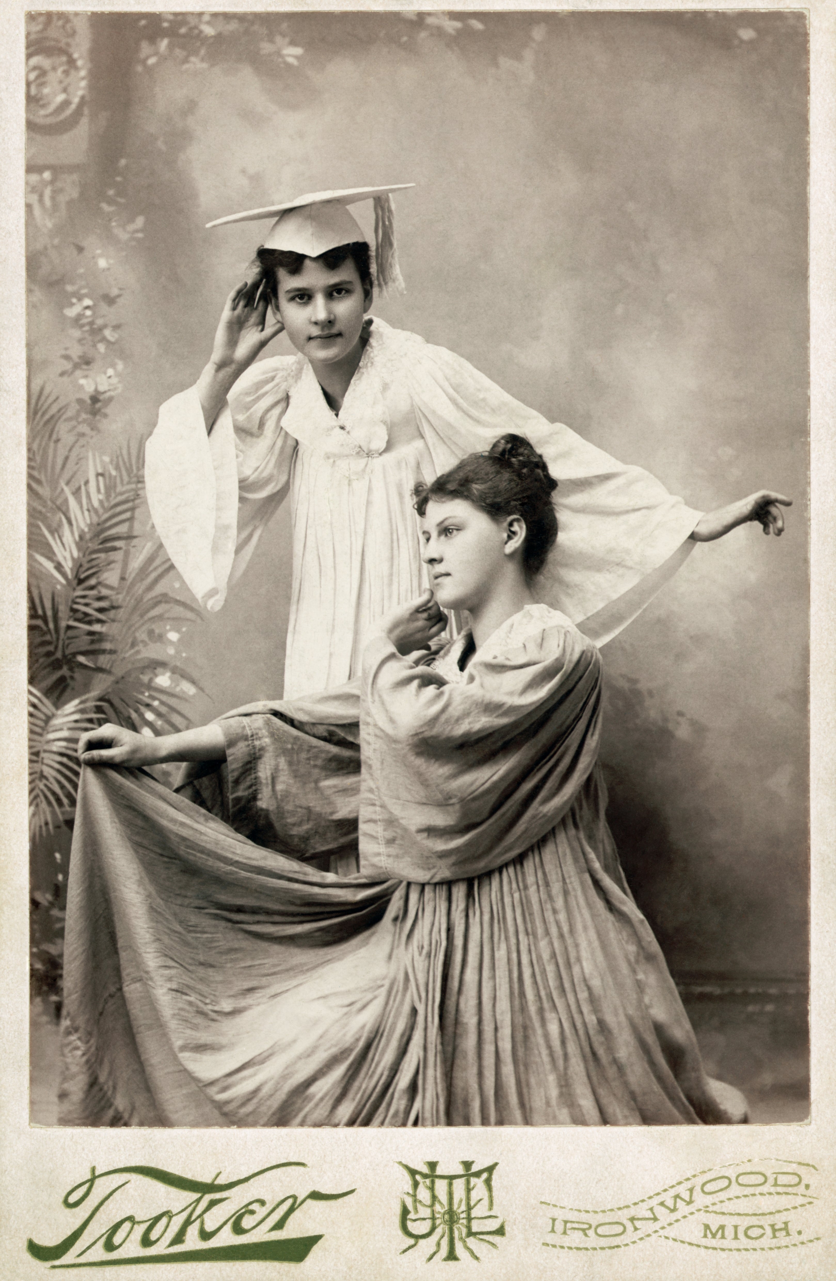 Two women in vintage attire; one wears a graduation cap and robe, looking pensive, while the other is seated with a long flowing dress