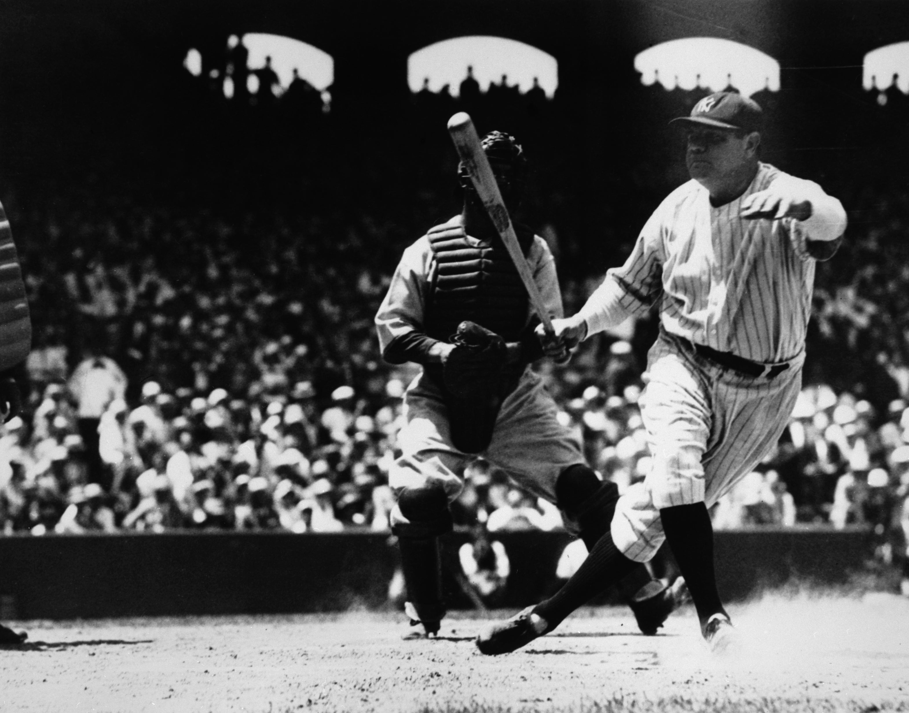 Baseball player Babe Ruth swings at a ball during a game, with a catcher and crowd in the background