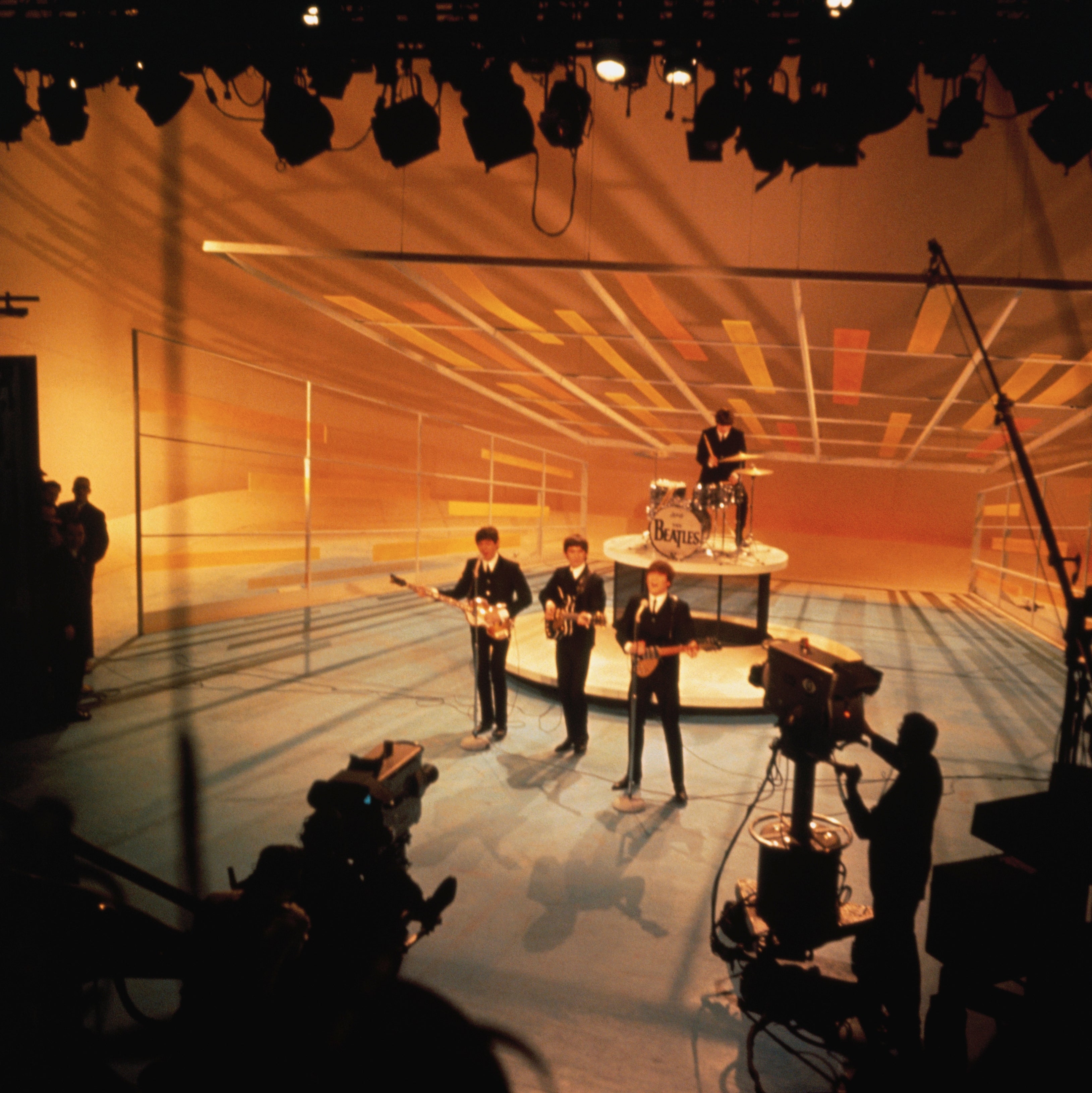The Beatles perform on a TV stage, three with guitars and one on drums. Stage hands and cameras surround the set