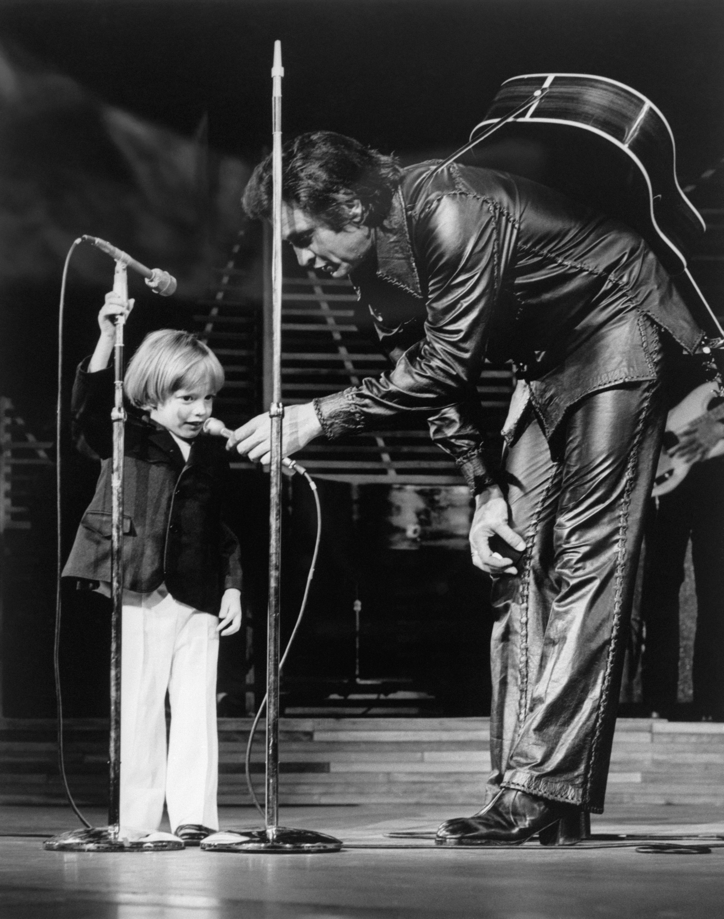 Johnny Cash in a leather outfit holds a microphone for his child, John Carter Cash, on stage. The child is reaching up to speak or sing into the mic
