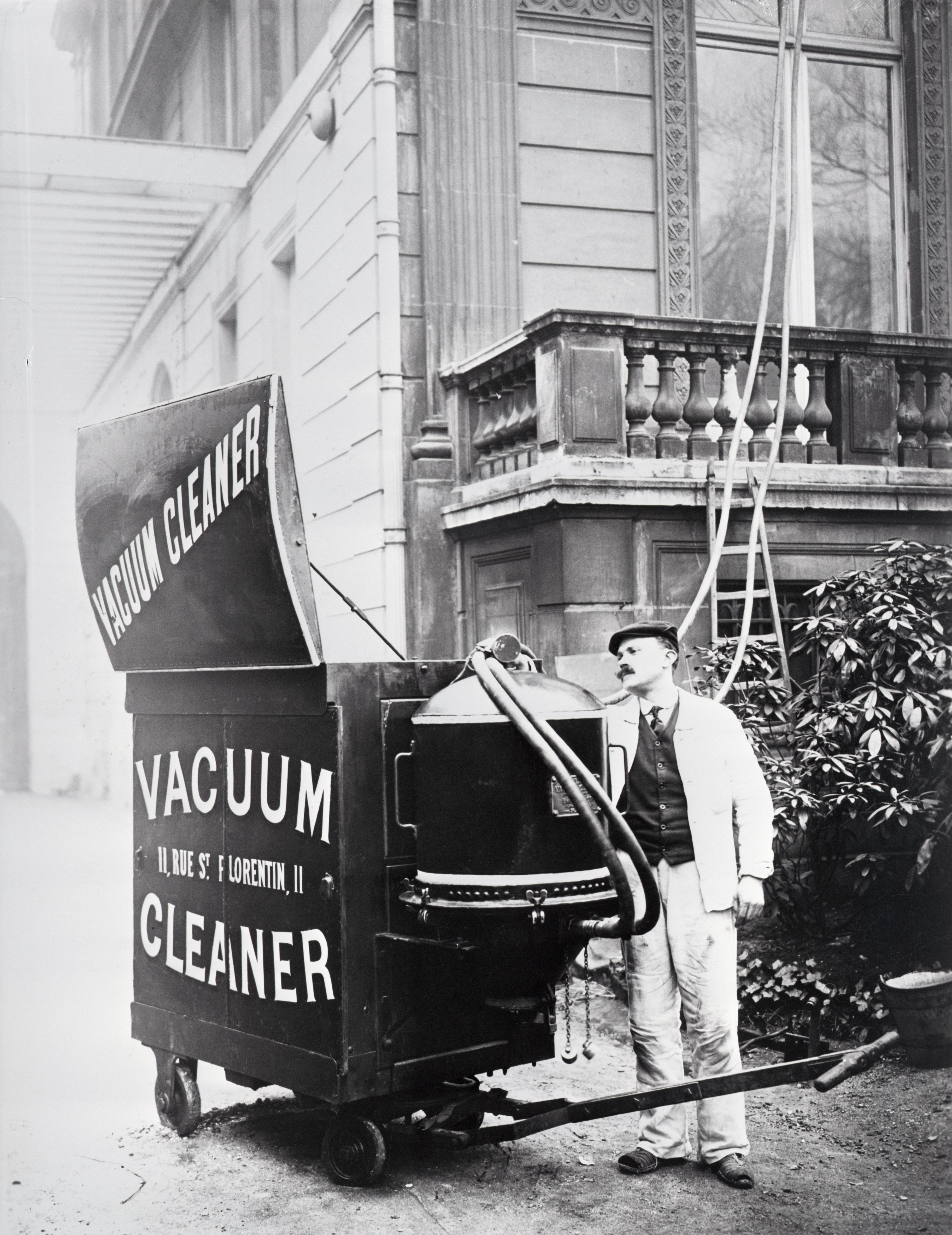 A man operates a large, early 20th-century street vacuum cleaner with "Vacuum Cleaner" printed on it outside a building