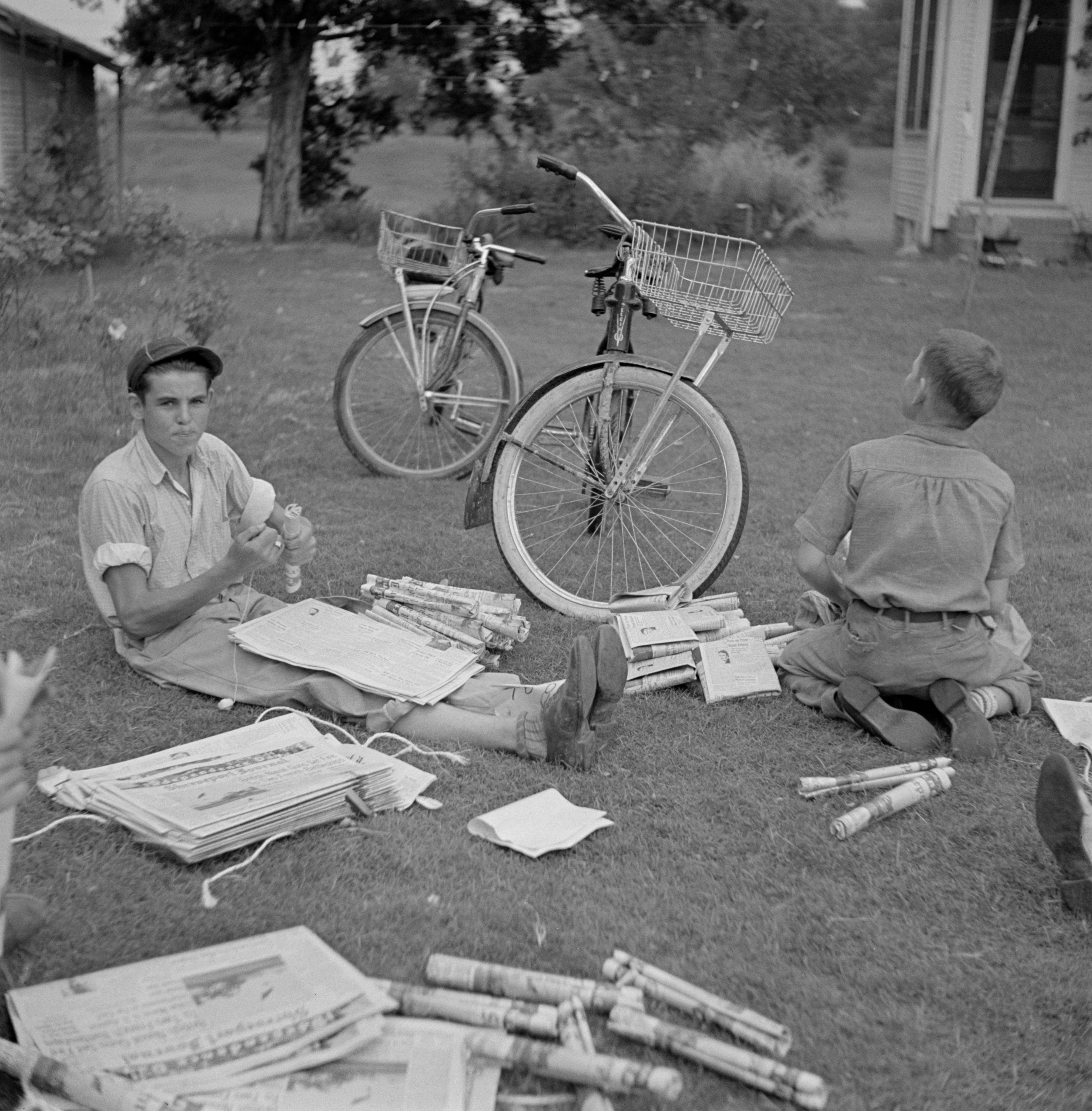 Children seated on grass, folding newspapers near bikes. A lawn and a house are in the background