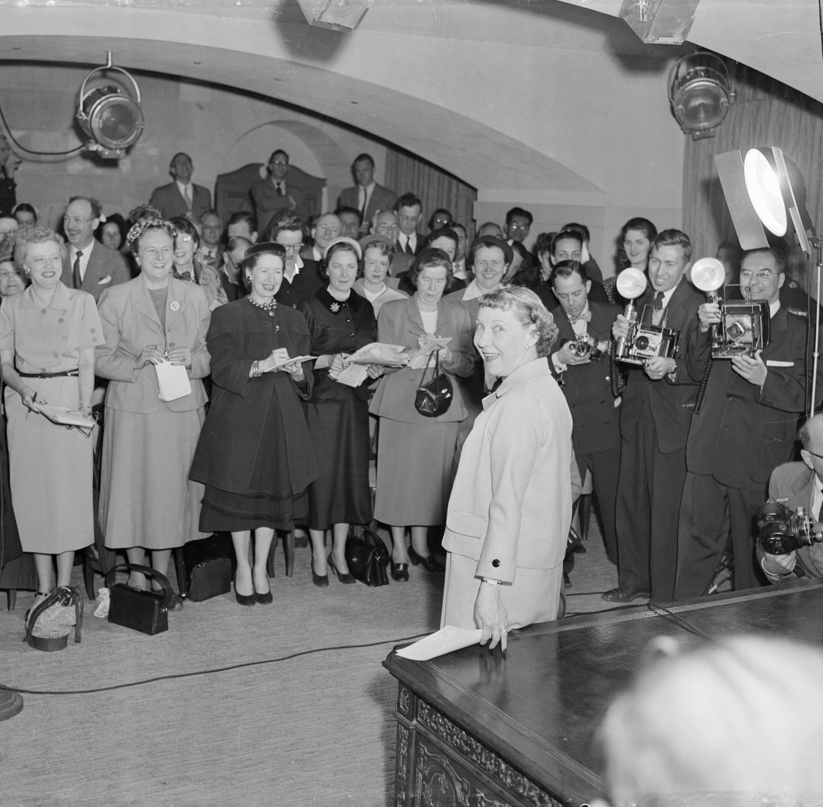 Mamie Eisenhower stands confidently at the foreground, facing a group of reporters and photographers in a busy press room