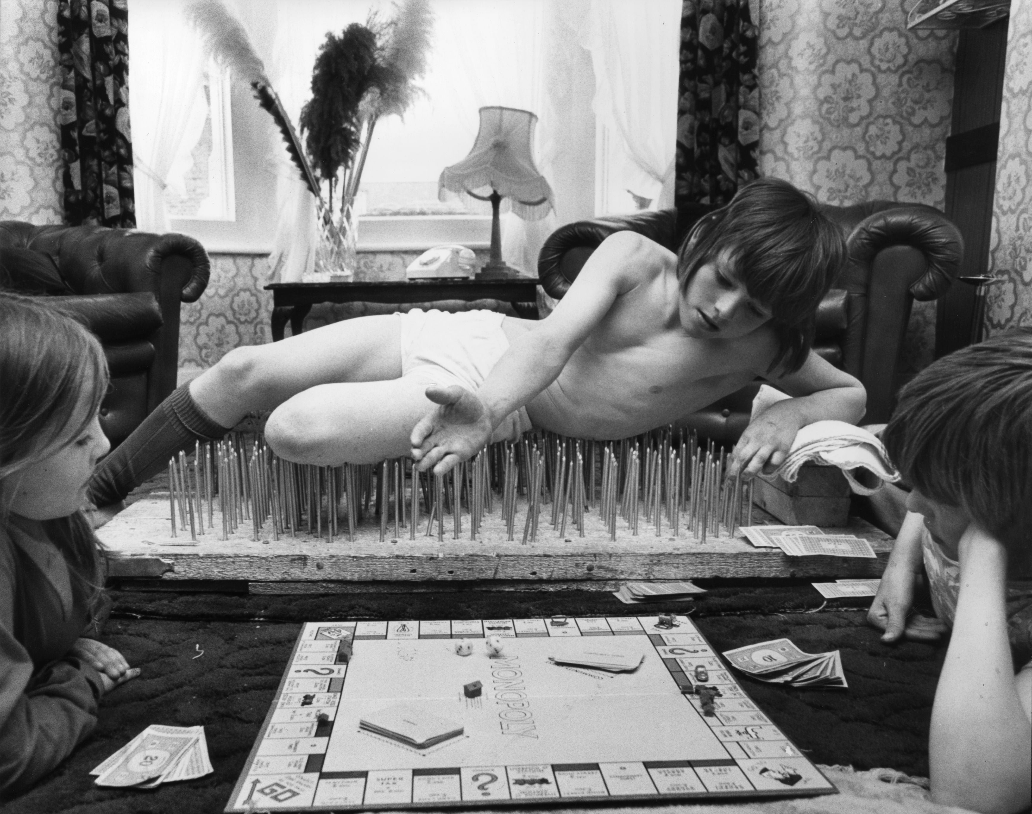 A young person dressed casually lies on a bed of nails; two others watch nearby while the Monopoly board game is set up in front of them