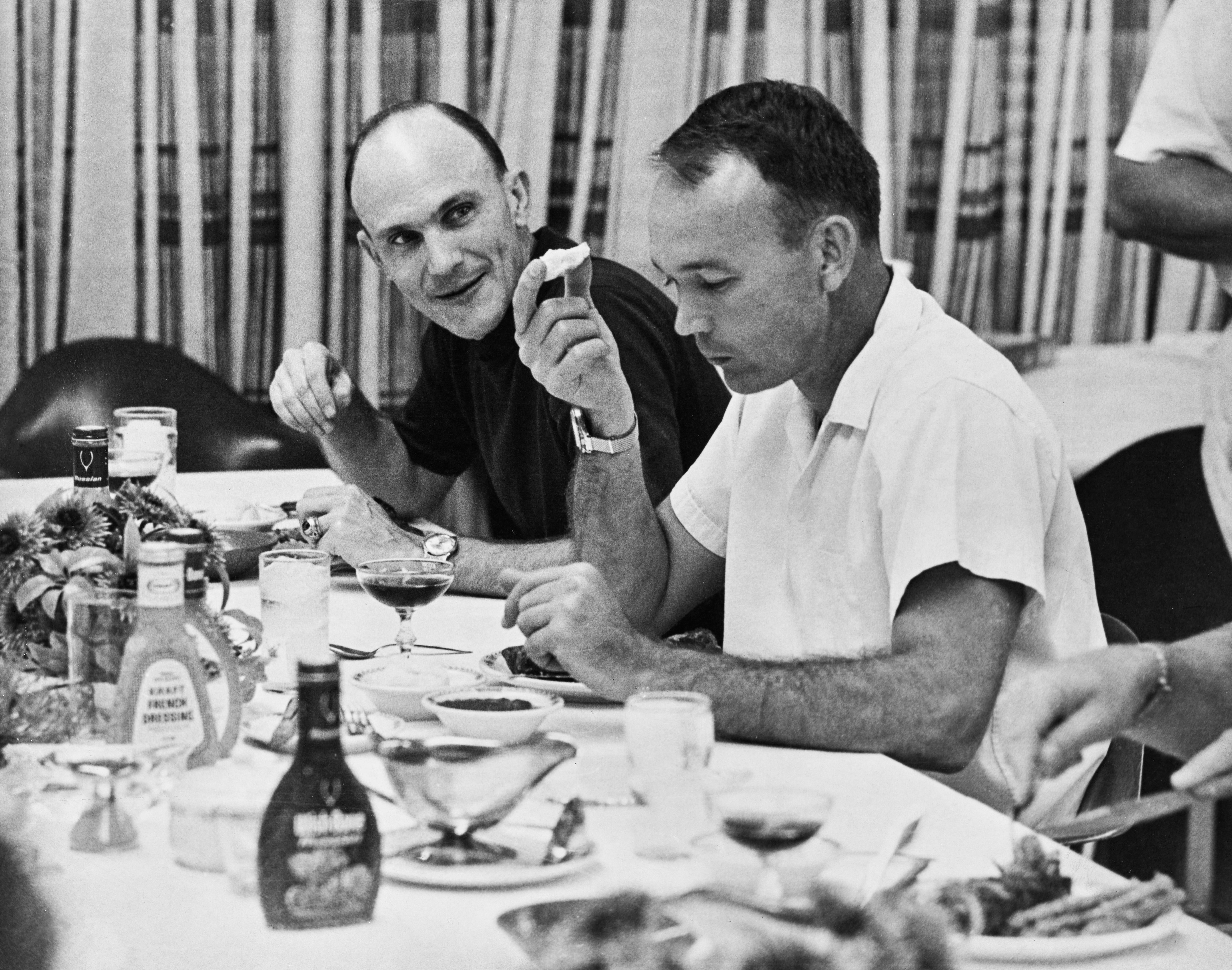 Ken Mattingly and Michael Collins seated at a dining table with various food and dressing bottles