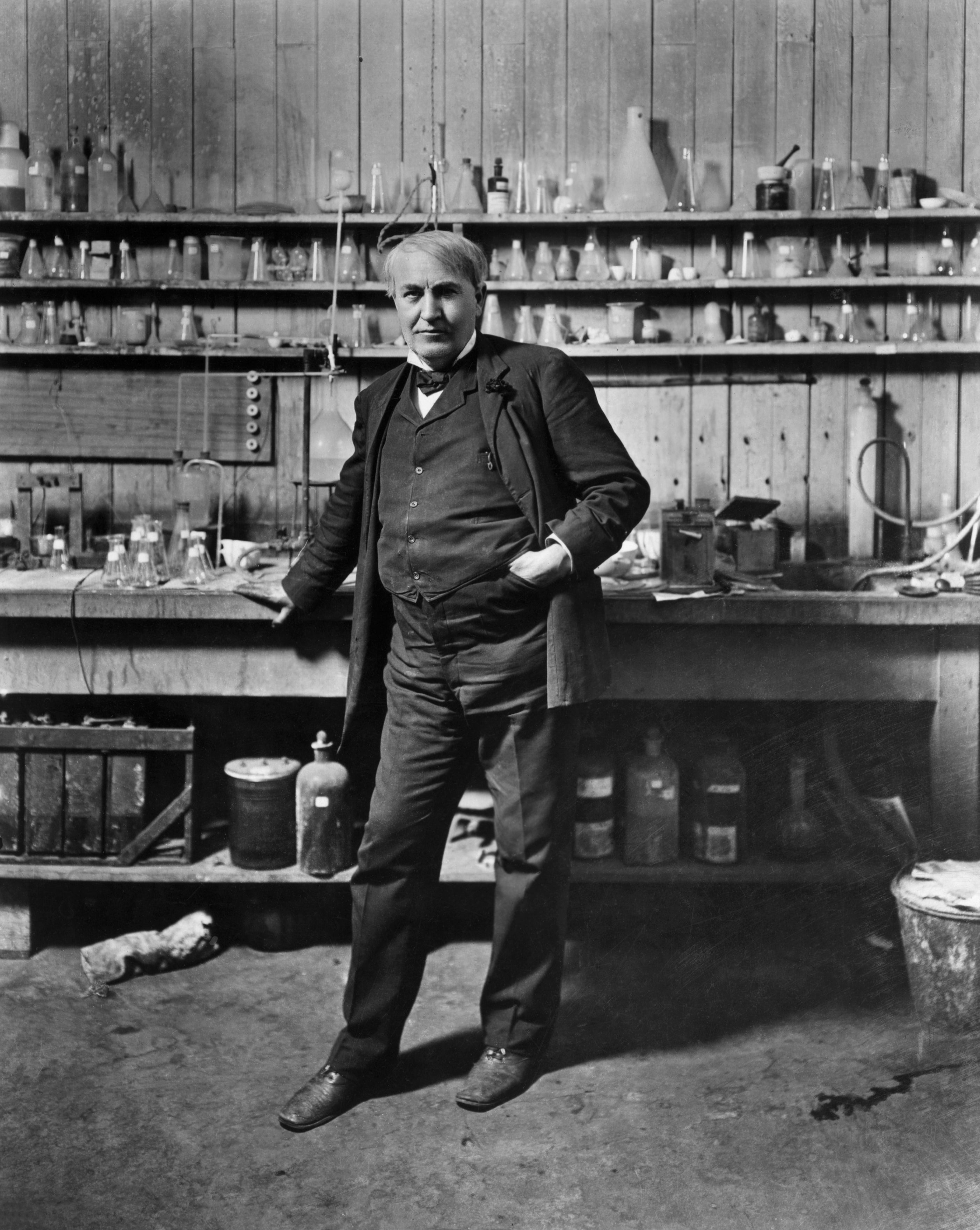Thomas Edison in historical attire stands confidently in a laboratory filled with bottles and equipment