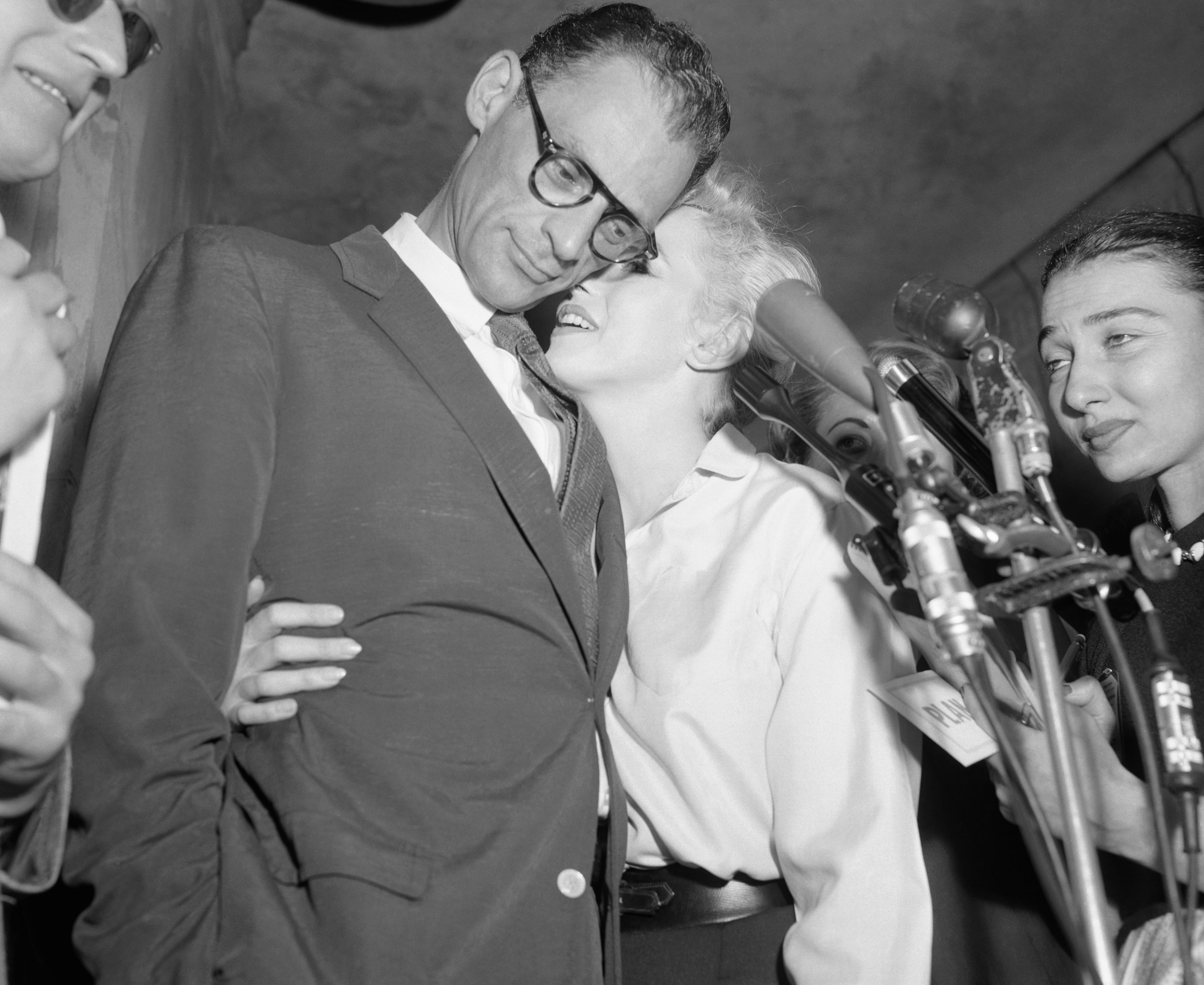 Marilyn Monroe in a blouse leans affectionately towards Arthur Miller in a suit and glasses at a press event, surrounded by microphones and people
