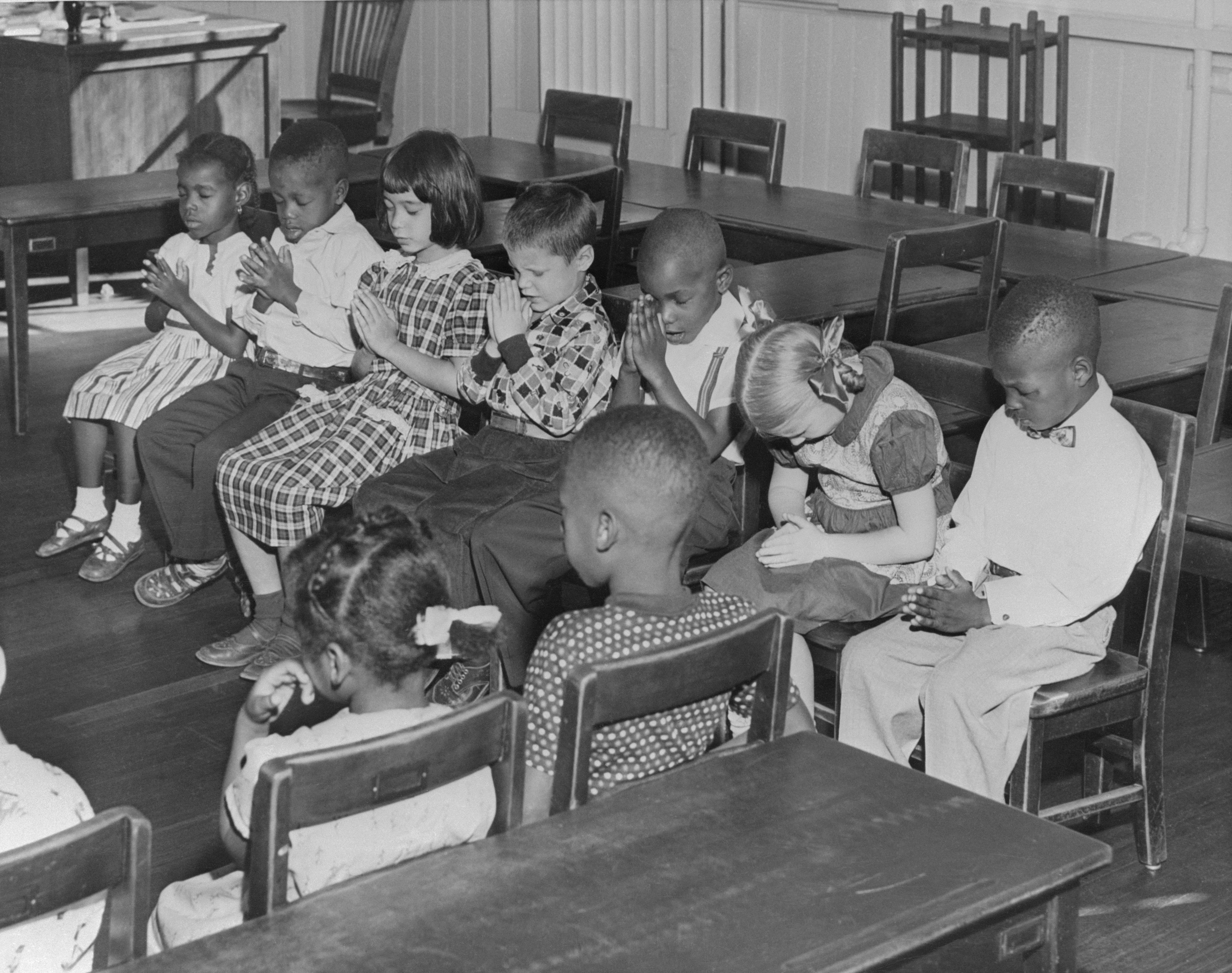 Children sitting in a classroom, some with heads bowed, praying. The room has desks and chairs arranged in rows